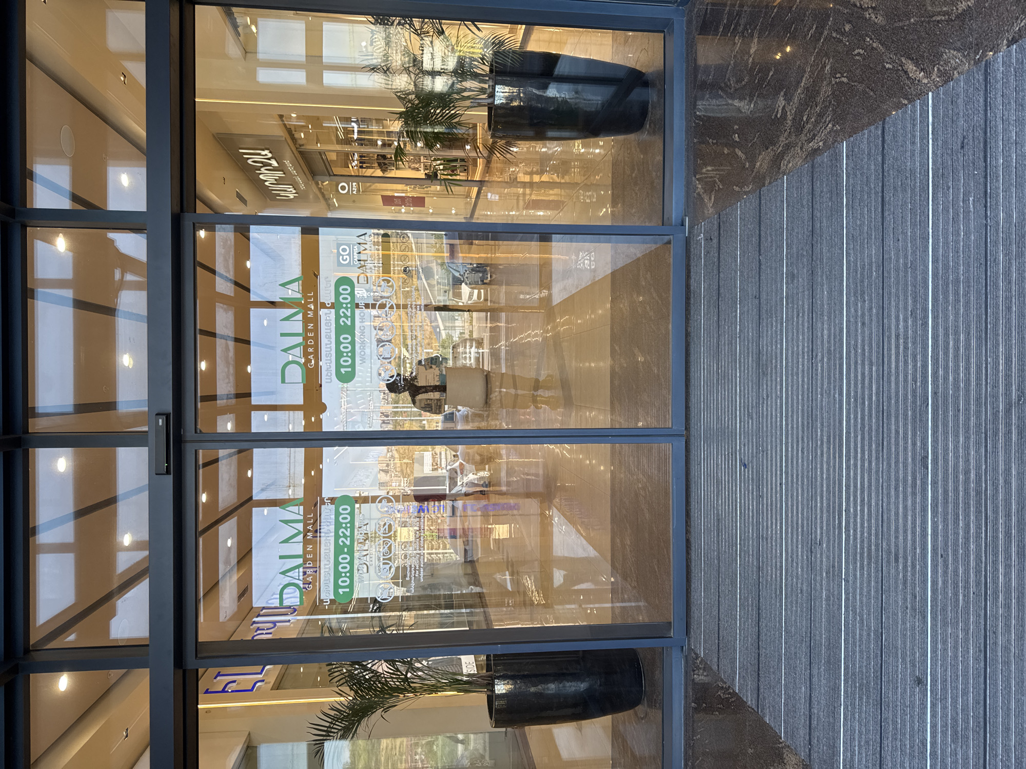 This is a photograph of the entrance to the Dalma Garden Mall in Yerevan, Armenia, as viewed from the exterior. The entrance consists of large glass sliding doors with dark gray framing. On the glass doors, there are green and white signs displaying the mall's name, "DALMA GARDEN MALL," and its operating hours: "10:00-22:00." There are also Armenian inscriptions on the signs.
To the left of the entrance, a tall, black, cylindrical planter holds a green palm plant. To the right, a similar planter with a palm plant is visible through the glass. The floor in front of the entrance is covered with a dark gray mat with horizontal white stripes. The floor inside the mall is shiny and reflective, showing the overhead lighting and the architecture of the interior.
Reflected in the glass doors are people, cars, and the surrounding environment, suggesting an outdoor urban setting. One person is seen walking through the entrance, wearing a backpack. The lighting in the scene is bright, indicating it is daytime. The overall impression is of a modern shopping mall entrance.