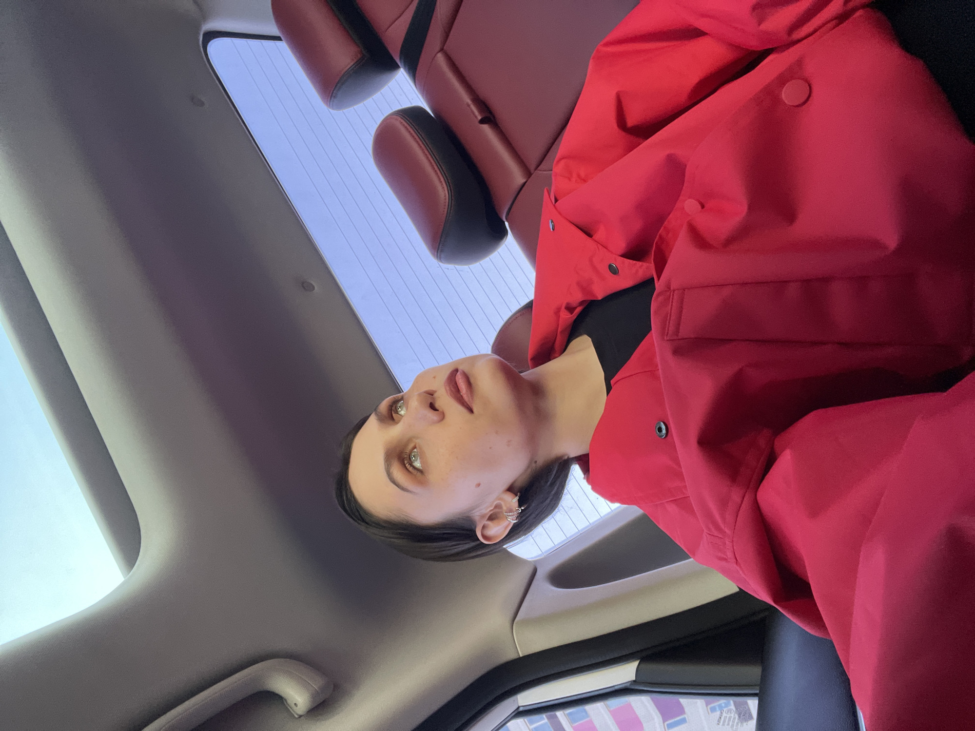 A woman is visible in the back seat of a car. She is wearing a red jacket over a black top. The car's interior is gray, with red leather seats. Outside the car window, a building is visible, suggesting an urban environment. The location is Izhevsk, Russia.