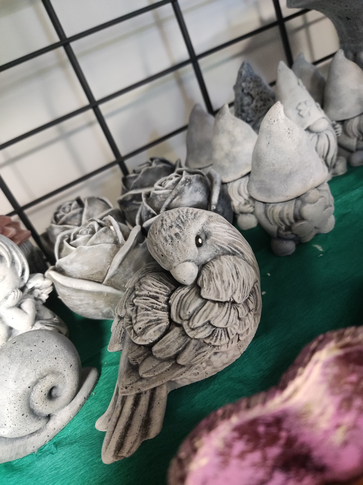 A close-up, eye-level shot shows a collection of grey concrete garden ornaments, including gnomes with pointed hats, roses, a snail, and a bird with ruffled feathers, all arranged on a green surface. In the background, a black metal grid is visible. The scene appears to be indoors, possibly in a shop or home. There is no indication of a specific location within the city of Kryvyi Rih, Ukraine.