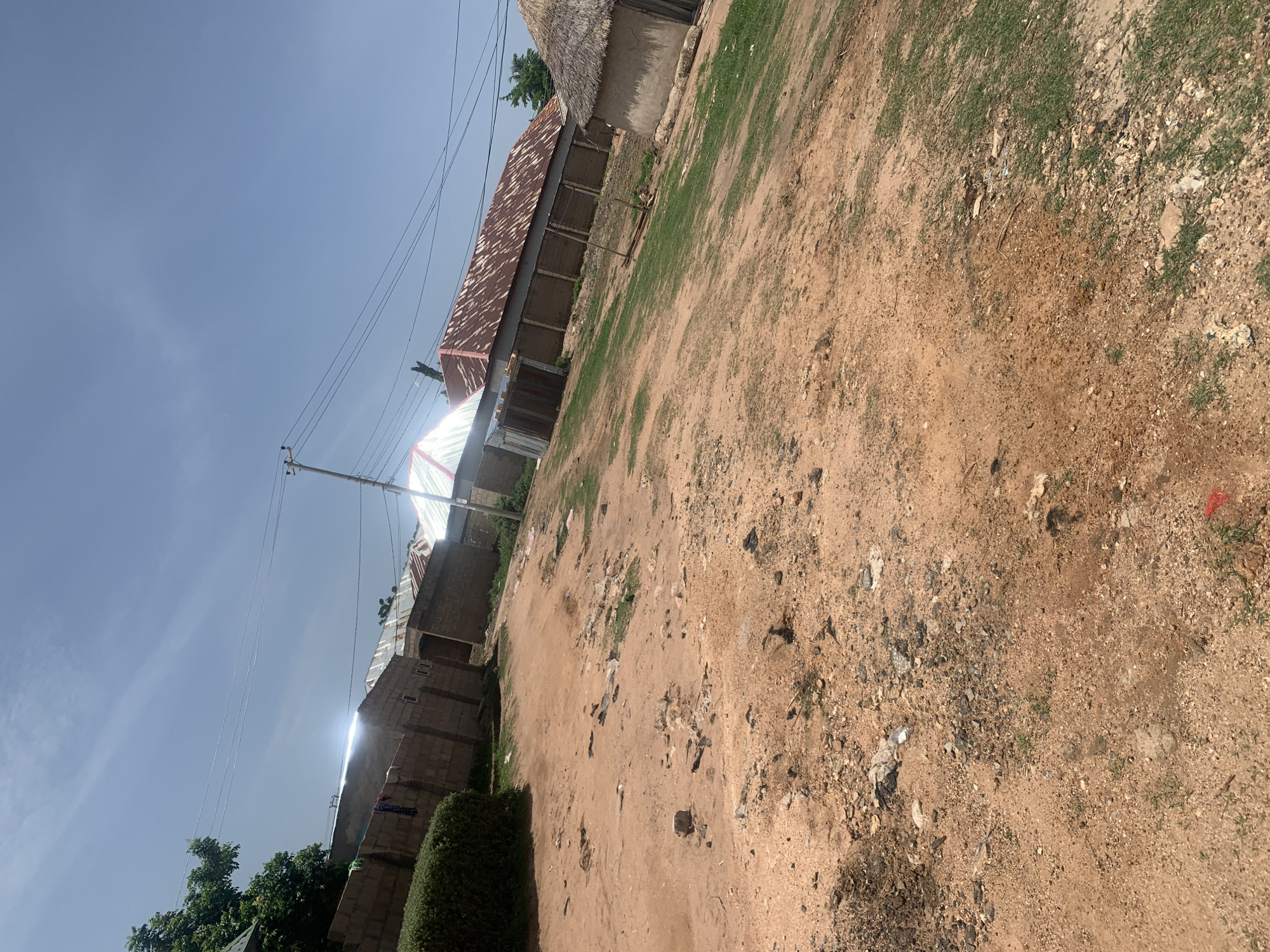 The image shows a dirt road with sparse vegetation and several buildings in Jauro Yinu, Nigeria. The buildings have various roof types, including corrugated metal and thatched. Some of the structures appear to be made of concrete blocks. The ground is sandy and uneven, with scattered debris. A power pole with electrical wires is visible in the background, indicating the presence of electricity infrastructure. The sky is clear with a few wispy clouds. The scene suggests a rural or semi-urban environment.