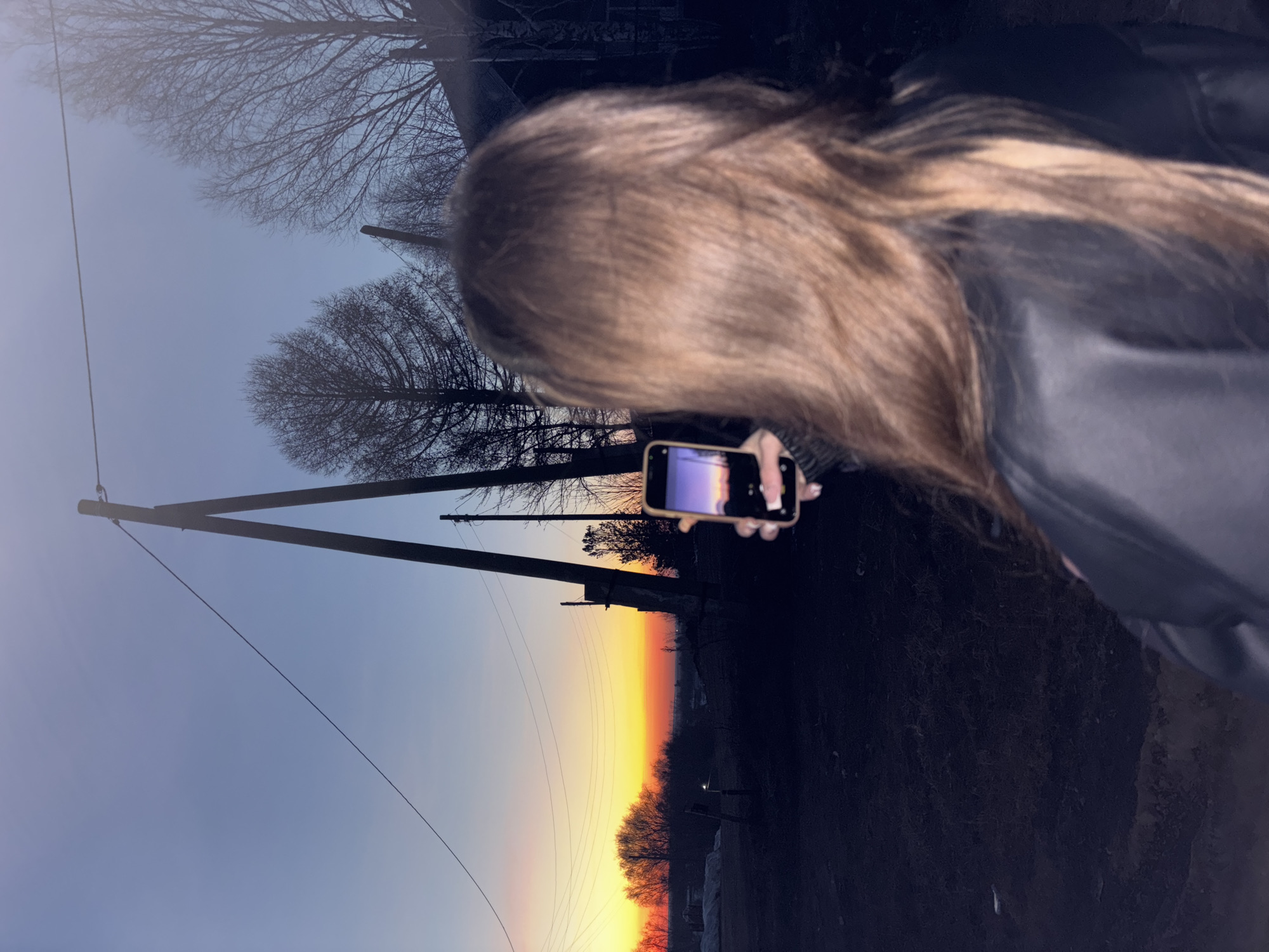 A woman with long brown hair is holding a smartphone and photographing a sunset over a road. Utility poles and bare trees are visible along the road. The location is possibly in or near Фоки, Russia, indicated by similar landscapes and infrastructure in online images. The sky transitions from orange to blue, indicating the time of day. The woman is wearing a dark jacket.