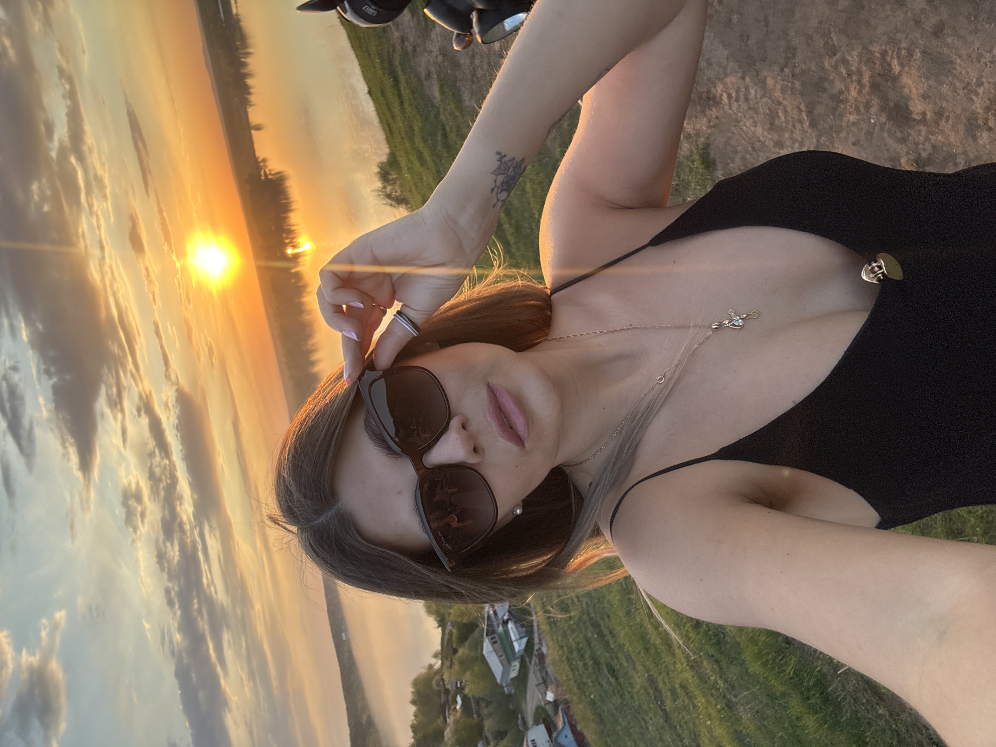 A selfie-style photograph depicts a woman standing outdoors at sunset, possibly in the vicinity of Ольховка, Russia, given the visible landscape with a body of water. The woman, centered in the frame, is wearing a black low-cut top and sunglasses, which she appears to be adjusting. She has brown hair and fair skin, with a tattoo visible on her arm. A delicate necklace adorns her neck. In the background, the setting sun casts a warm glow over a body of water reflecting the sky, with some vegetation and what appears to be a village visible in the distance.