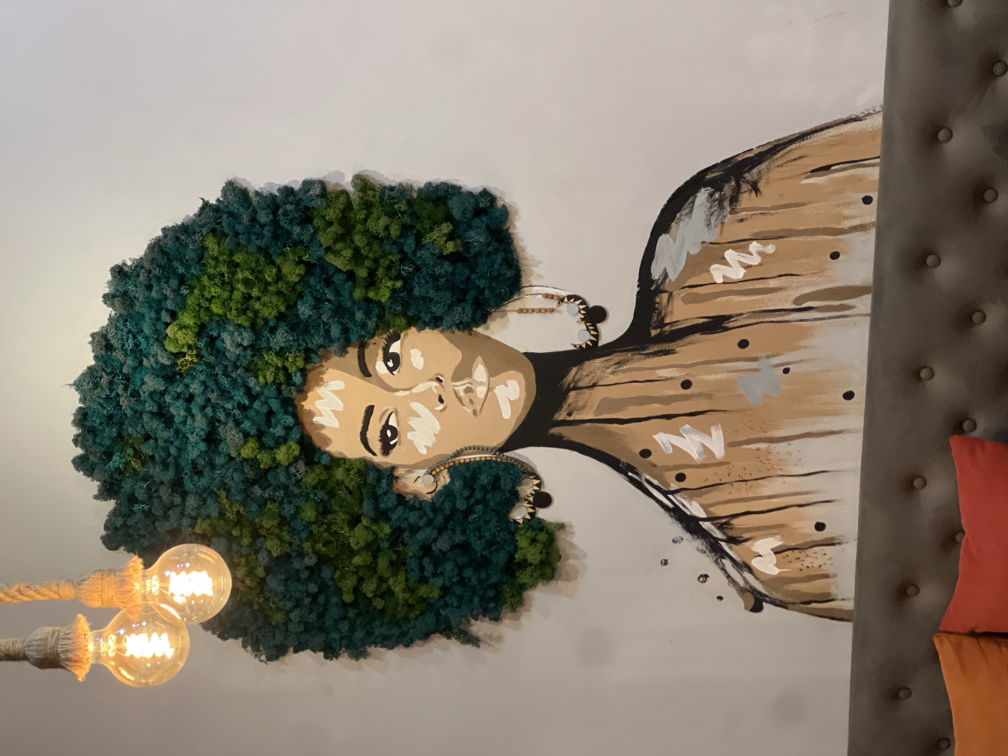 The image shows a wall decoration featuring a portrait of a Black woman with an afro made of green and blue moss. Her skin is painted in shades of brown with white highlights, and she has large earrings. The artwork is mounted on a white wall. To the left, a lamp with two exposed bulbs hangs from a rope. To the right, a grey upholstered headboard with tufted buttons is visible, along with two colorful pillows. The image was taken indoors in Yerevan, Armenia.