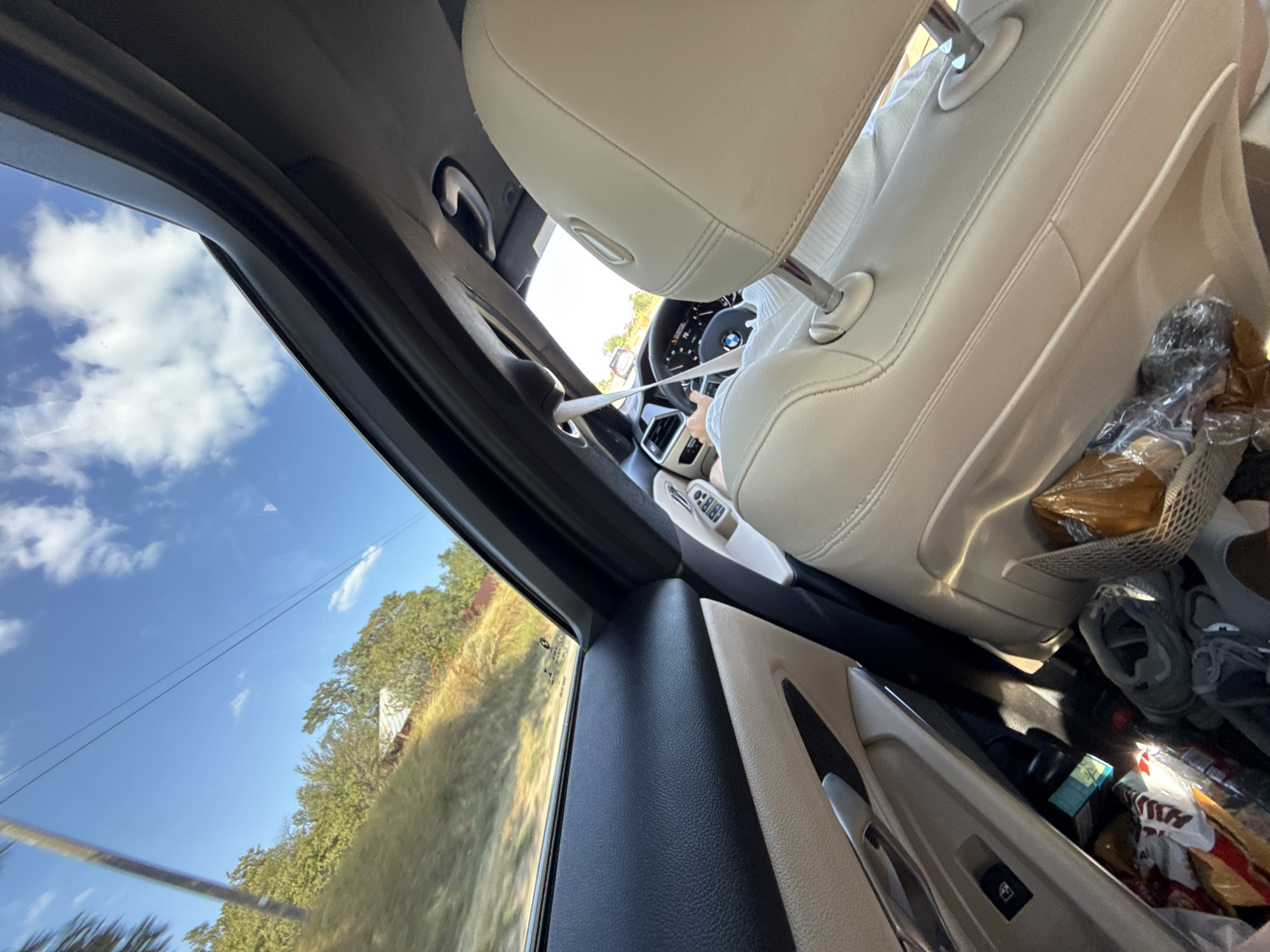 The image depicts the interior of a car, looking out through the sunroof and side window. The car appears to be in motion, with a landscape of trees, a grassy hill, and a road visible outside. The interior features light-colored leather seats, a steering wheel, and dashboard instruments. There are also several bags and items scattered on the floor of the car. The sky is blue with scattered white clouds. The location is identified as being in Lehancea, Romania.