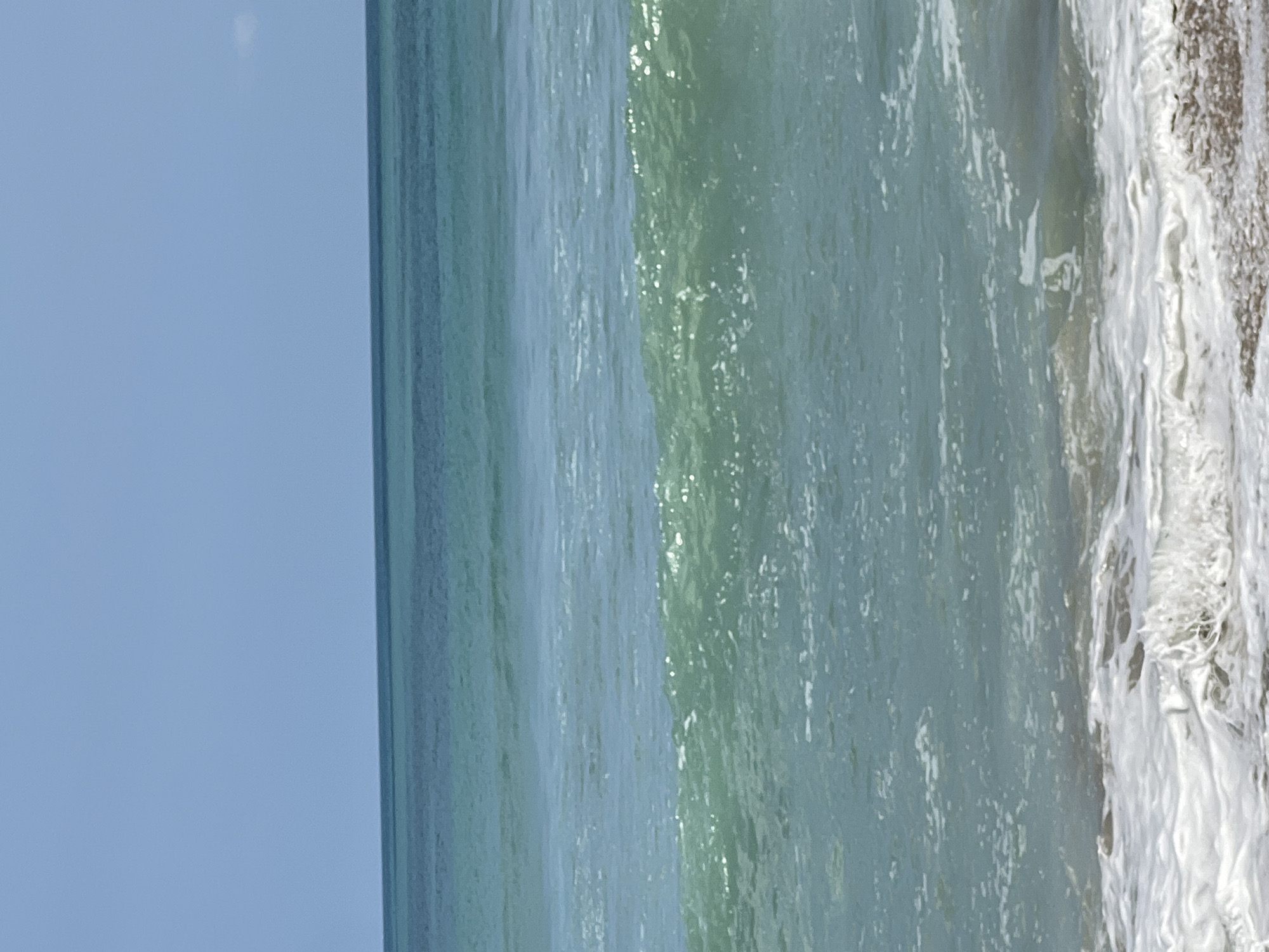 A serene ocean scene unfolds with the clear blue sky meeting the tranquil turquoise waters of Kuta, Indonesia. Gentle waves ripple across the surface, with the closest wave cresting and breaking into white foam near the shore. The water transitions from a lighter, more translucent hue near the shore to a deeper, richer blue as it extends towards the horizon. The beach is a blur of pebbles and sand, hinting at the edge of the land. The image is devoid of people, vehicles, or man-made structures, presenting an untouched natural landscape. The lighting suggests a bright, sunny day, casting a soft glow over the water.