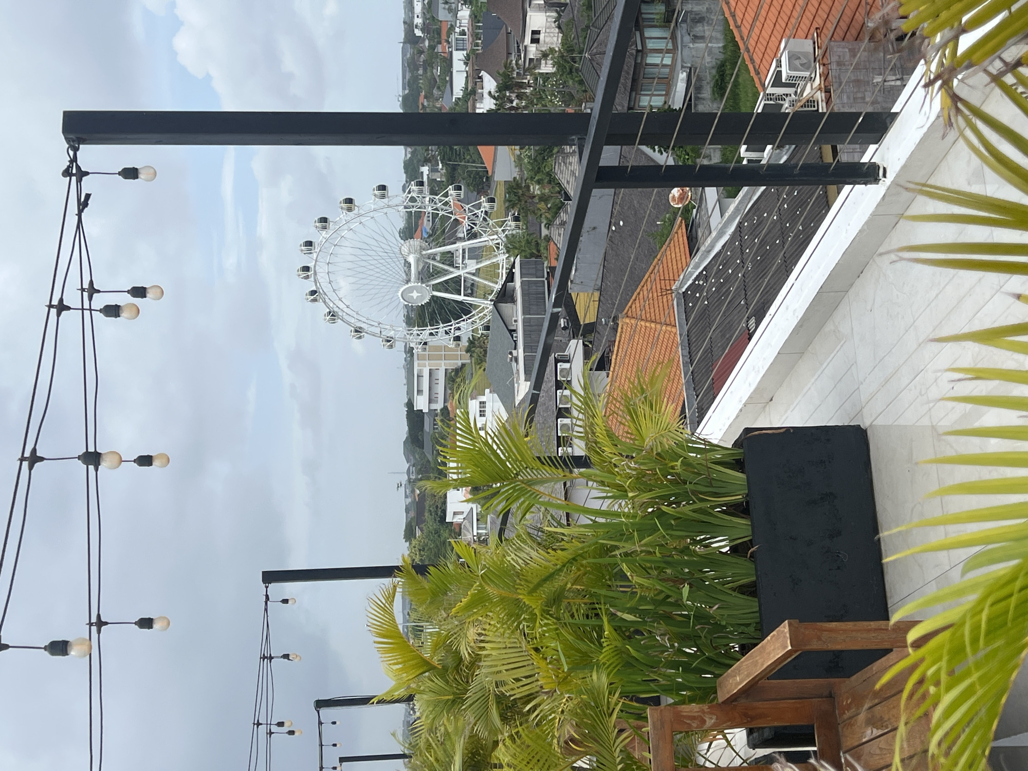 The image depicts a view from a rooftop in North Kuta, Indonesia, overlooking the city. The most prominent object in the distance is a large white Ferris wheel. The foreground features outdoor lighting strung along black metal poles. Palm trees are planted in black planters on the rooftop. Buildings with a mix of tiled and flat roofs are visible in the midground. The sky is overcast.