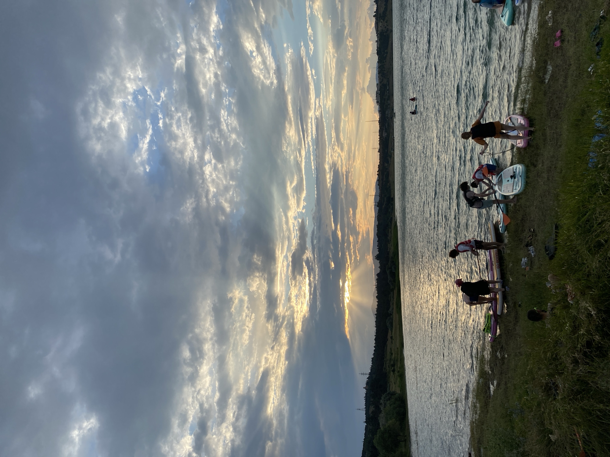 This is a vertical, medium shot of a lake scene at dusk, with a focus on people preparing to paddleboard. The sky is filled with dramatic, textured clouds, with rays of sunlight breaking through them near the horizon, casting a golden glow. In the foreground, several individuals are gathered on the grassy shore next to a collection of paddleboards. Some are standing and interacting with the boards, while others appear to be getting ready to launch. The lake itself is rippling, reflecting the muted colors of the sky. In the distance, across the water, is a treed shoreline with a few distant structures and power lines, suggesting a semi-urban or suburban environment. The scene evokes a sense of outdoor recreation and natural beauty at the end of the day. The lake is likely Lisi Lake in Tbilisi, Georgia, a popular spot for water activities.