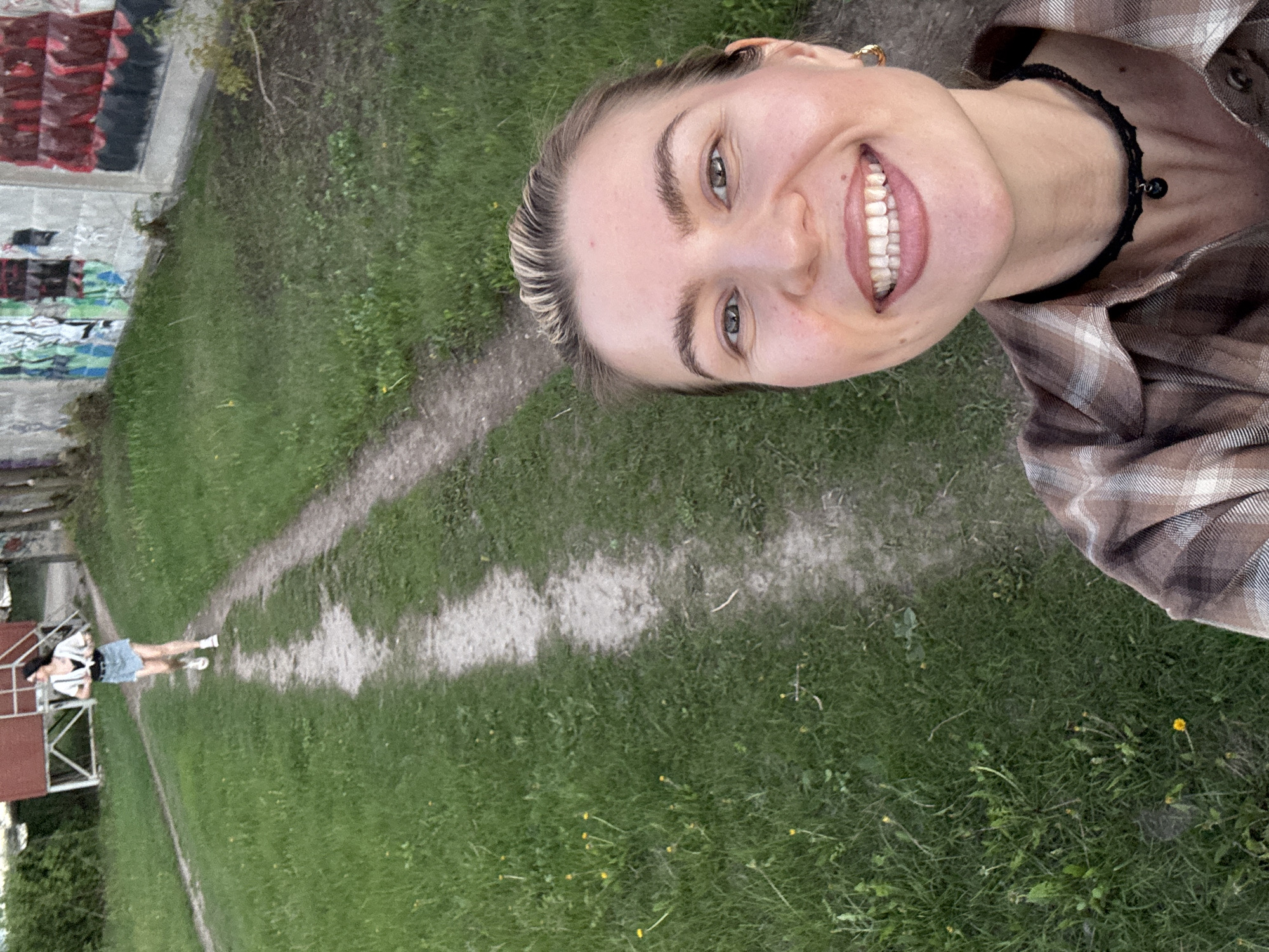 The image is a selfie of a fair-skinned person with blonde hair, smiling, and wearing a brown plaid shirt and a black beaded necklace. The person is standing in a grassy area with a dirt path, possibly a park. In the background to the left, there is a building with graffiti on it and a figure of a woman standing near a red structure. The location is unknown, as are the identities of the people. The environment consists of green grass, a dirt path, and structures that suggest an urban or semi-urban area.