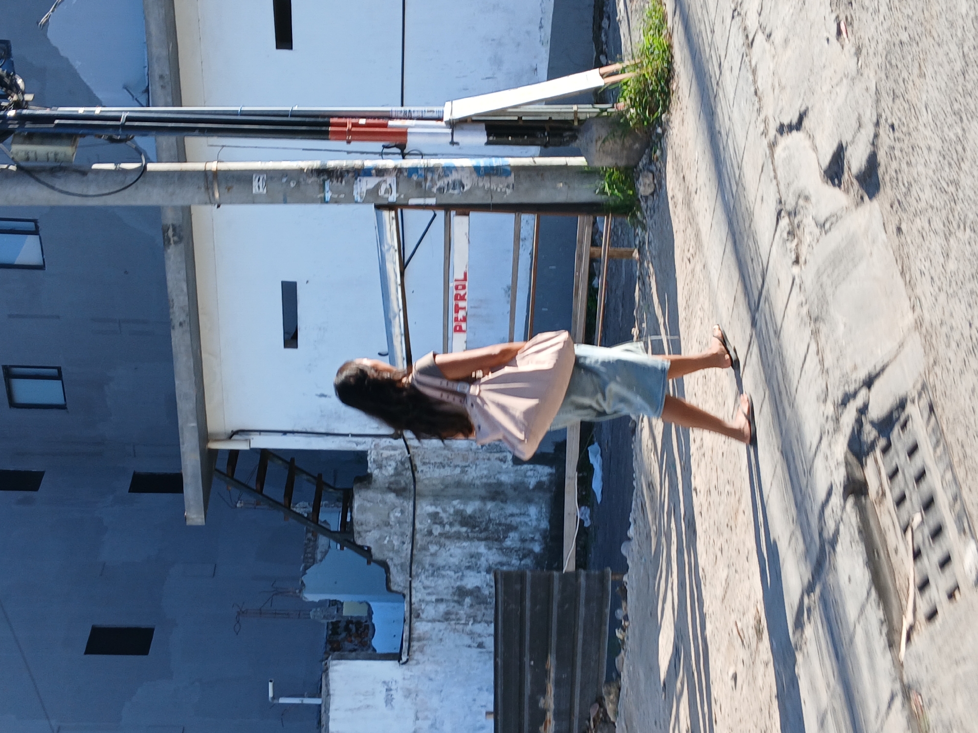 In North Kuta, Indonesia, a woman with dark, long hair, seen from the rear, walks on a paved sidewalk. She is wearing a light-colored short-sleeved shirt and a denim skirt, with flip-flops on her feet. She carries a large, light pink shoulder bag across her right shoulder. Behind her, a metal fence with "PETROL" written in red lettering is visible. The environment features a partially constructed building with a blue facade and multiple windows, adjacent to a white structure. A metal staircase is attached to the side of the blue building. A utility pole with wires stands near the fence. The ground is a mix of dirt and cracked pavement, with some vegetation growing near the base of the utility pole. Shadows suggest a bright, sunny day.