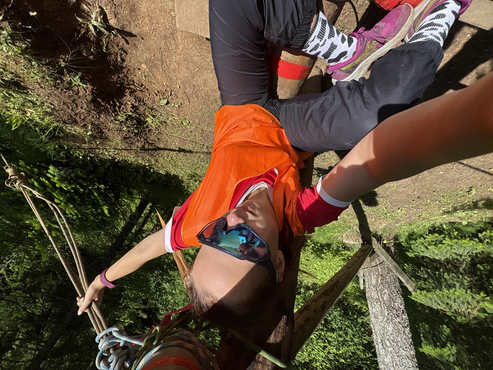 This is a first-person perspective photograph taken outdoors, likely on a sunny day given the strong shadows. The scene appears to be a forest or wooded area, with trees and vegetation visible.
The primary subject is a person, seen from the waist up, lying on their back with their head angled upwards towards the camera. They are wearing an orange vest over a red and white shirt, black pants, and distinctive black and white patterned socks with pink athletic shoes. They are also wearing large, mirrored sunglasses. A purple wristband is visible on their left wrist.
The person's arms are extended, with their left hand holding onto a bundle of thick ropes. These ropes, along with other lighter green and blue ropes, are attached to a wooden structure which appears to be part of some kind of adventure course or rigging. The person's legs are also visible, with one leg bent and the other extended. Red tape is wrapped around what appears to be a wooden support beam near their legs.
The background shows the earthy ground, grass, and the trunks and foliage of trees. The overall impression is of someone participating in an outdoor activity involving ropes and heights, possibly zip-lining, climbing, or a similar adventure sport. The lighting suggests it is daytime. There is no visible text in the image.