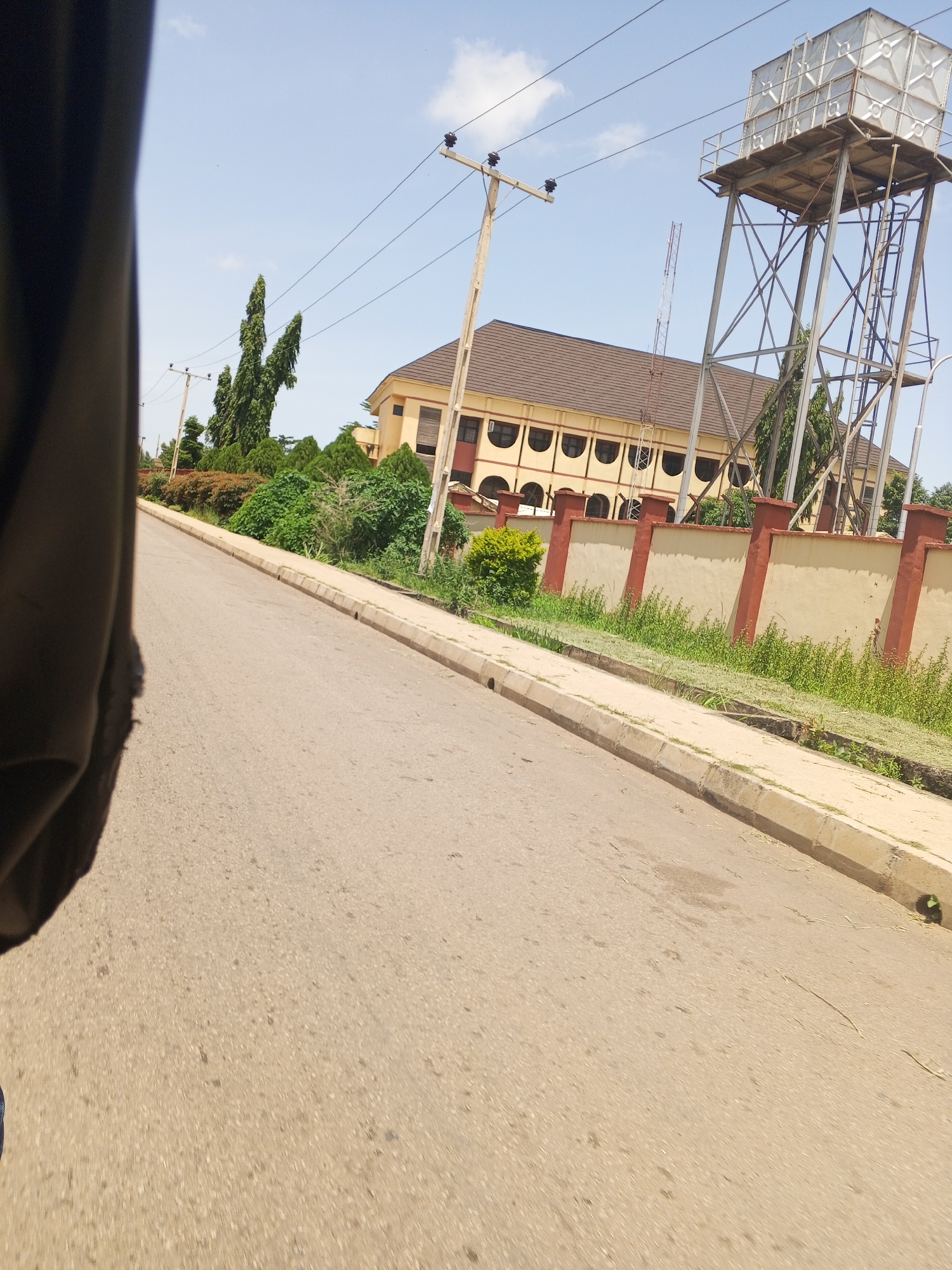 An exterior shot captures a street in New Stadium, Nigeria, on a sunny day with a bright blue sky. The road is paved and curves gently to the left. A concrete curb runs along the right side of the road, bordering a patch of grass and a low, cream-colored wall with decorative red pillars. Behind the wall, a light yellow, multi-story building with a brown roof is visible. The building features arched windows and a repeating pattern of them. To the right of the building, a tall, metal water tower stands, its structure supported by a network of beams. Power lines crisscross the sky, supported by wooden utility poles. Lush green trees and bushes are scattered throughout the scene, contributing to the urban landscape. The photo is taken from a low angle, possibly from a moving vehicle, as indicated by the blurry dark object on the left side of the frame, which appears to be part of a car interior.