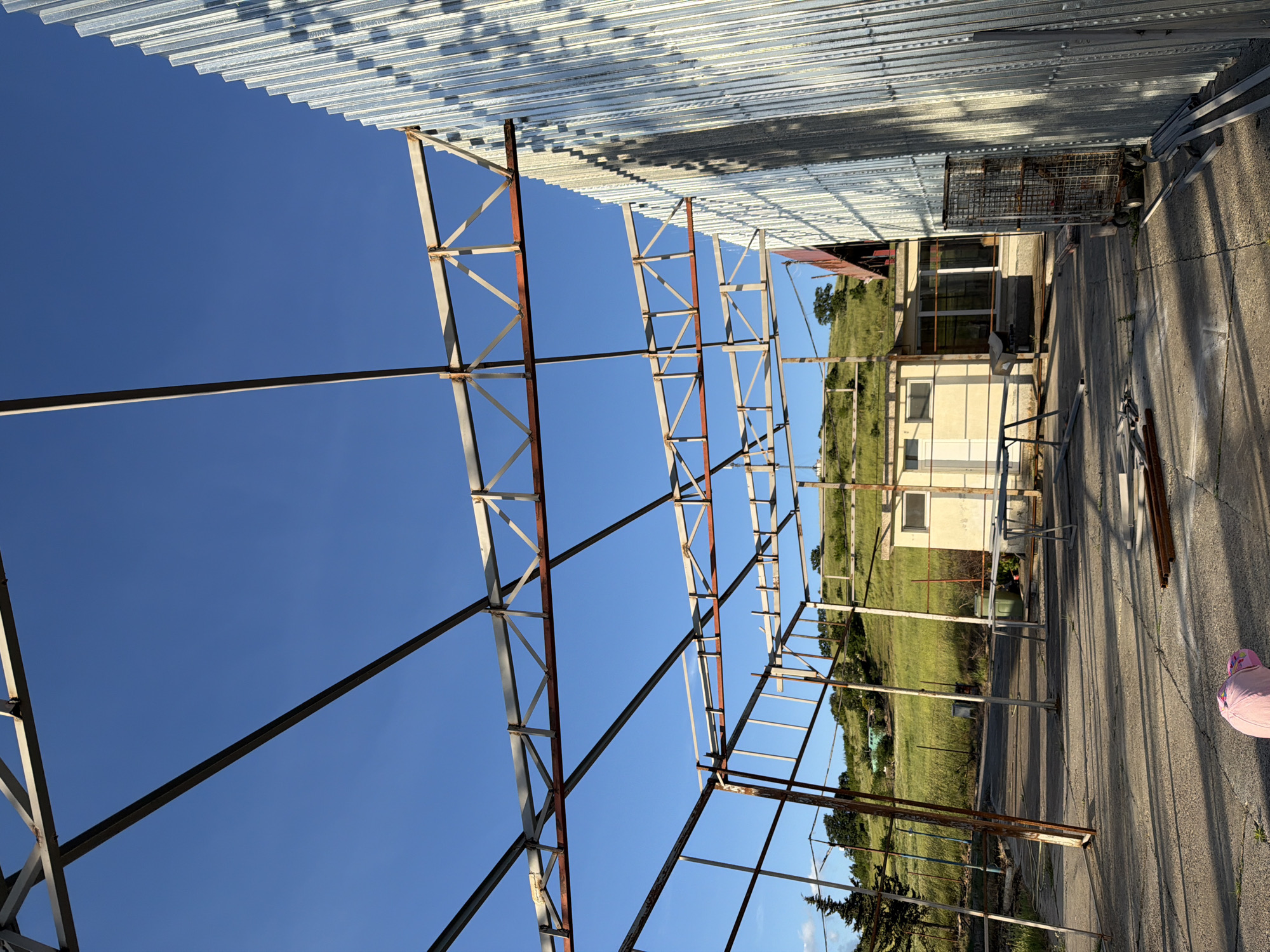The image shows a partially constructed metal framework against a clear blue sky in Dabnik, Bulgaria. The framework has a rusted appearance in sections. Above the framework is corrugated metal roofing. Beyond the framework, a grassy landscape slopes upward towards a building with light-colored walls and visible windows. On the paved ground adjacent to the framework, there is a metal shopping cart, metal pipes, and a pink object. The image is oriented such that the sky appears vertically in the middle.