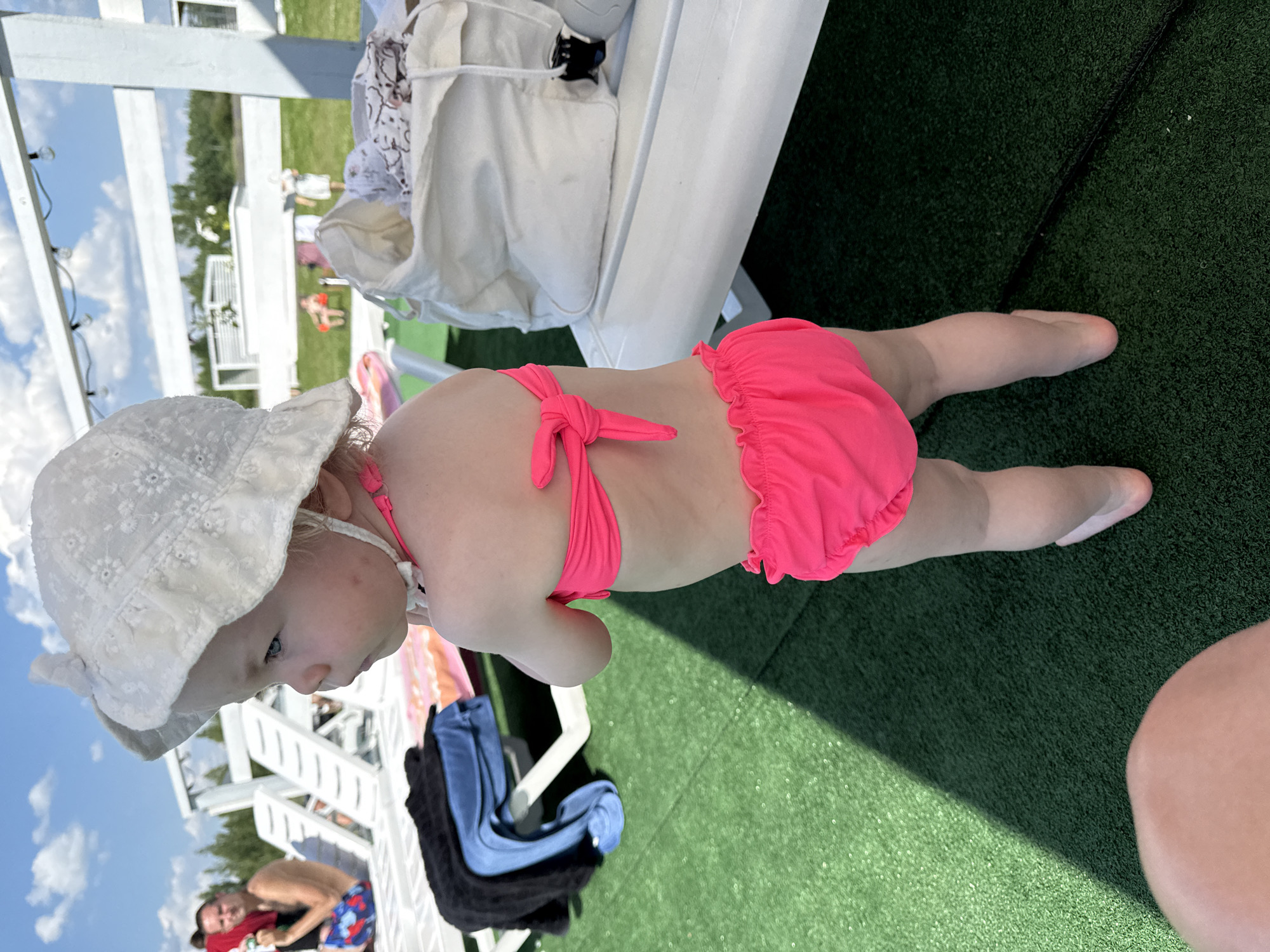A young child wearing a white sun hat and a bright pink bikini is lying on artificial turf next to white lounge chairs. The child's face is turned away from the camera, looking to the left. In the background, two adults are visible near lounge chairs, and the sky is bright blue with white clouds. One of the adults appears to be partially submerged in a body of water. The scene suggests a sunny day at an outdoor recreational area, possibly a pool or a resort, with people relaxing and enjoying leisure time. The artificial turf, lounge chairs, and distant trees indicate an outdoor setting. The time of day is likely daytime, given the bright sunlight casting shadows. The overall atmosphere appears relaxed and summery. There is no visible text in the image.