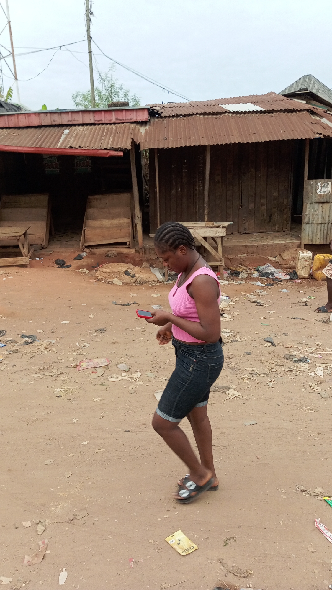 A woman walks on a dirt road in Onitsha, Nigeria, looking at a red cell phone she holds in her hands. She wears a pink tank top and dark denim shorts, with black sandals on her feet. The ground is covered in loose debris and trash. Behind her is a building with a rusted tin roof and wooden supports. The sky is overcast.