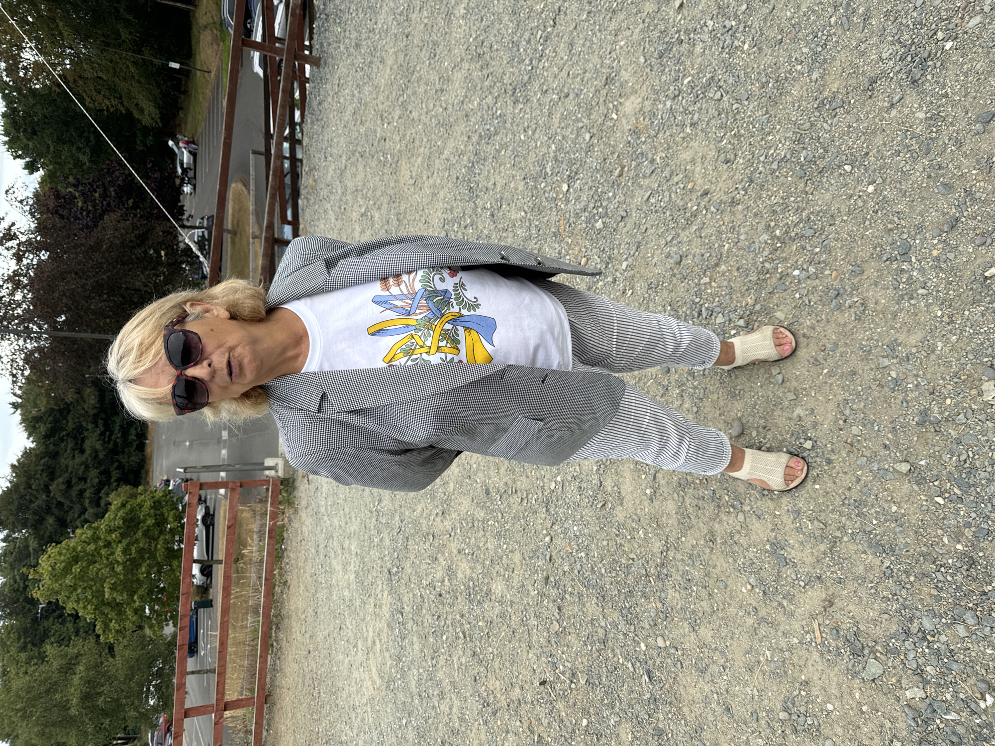 A full-body, eye-level shot shows a woman standing on a gravel surface in Courtown, Ireland. She is wearing a black and white patterned blazer over a white t-shirt adorned with a blue and yellow floral emblem. Her pants are striped in white and grey. The woman has blonde hair, wears large sunglasses, and is wearing light beige sandals. Behind her, a brown wooden fence lines the foreground. Beyond the fence, a paved parking lot is visible with several parked cars. Trees and foliage form a backdrop to the scene.