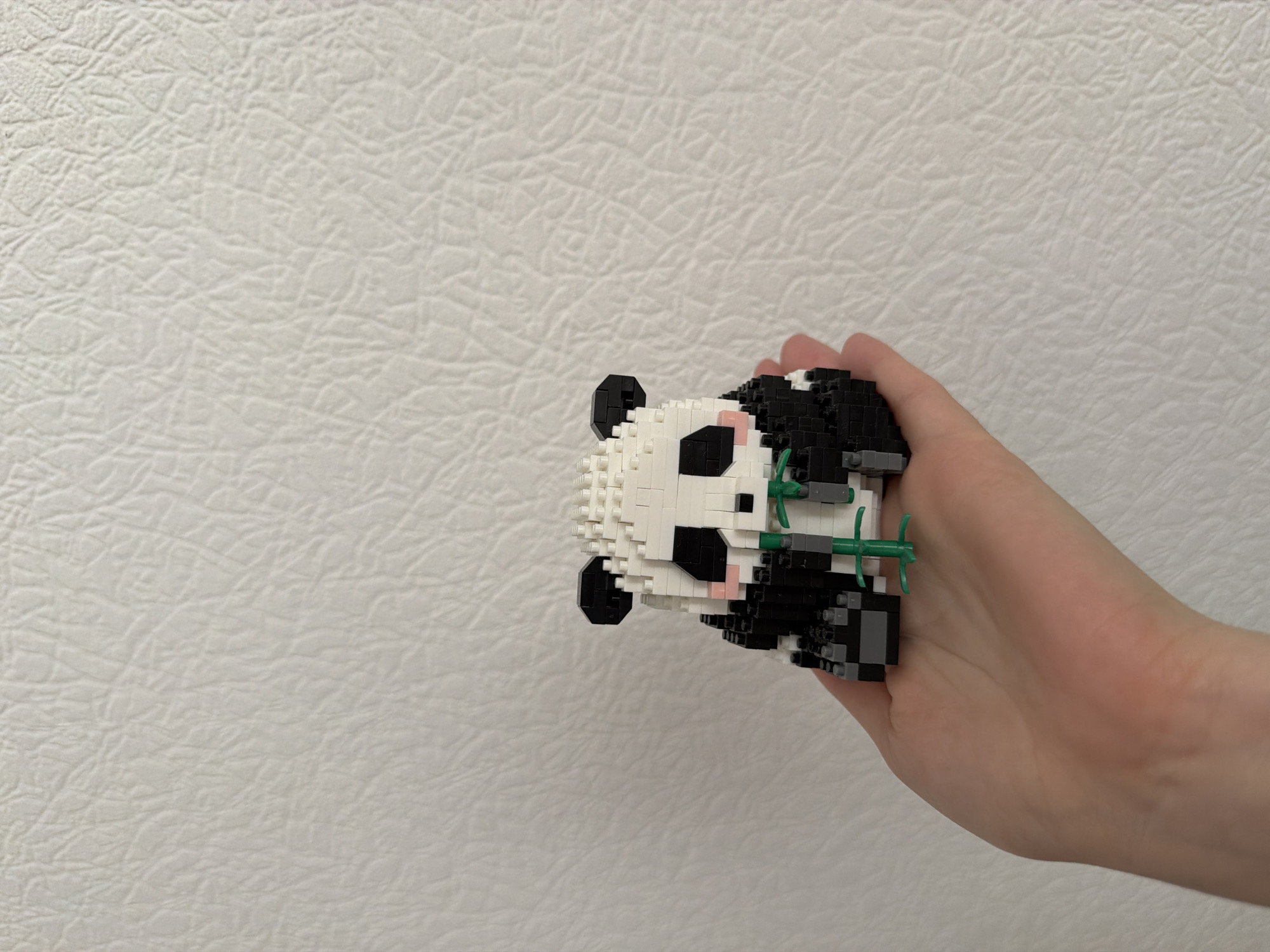 A close-up, eye-level shot shows a person's hand, palm up, holding a small, pixelated panda figurine made of white and black building blocks. The panda is sitting and holding a green bamboo stalk. The background is a textured white wall. The image was taken in Kyiv, Ukraine.