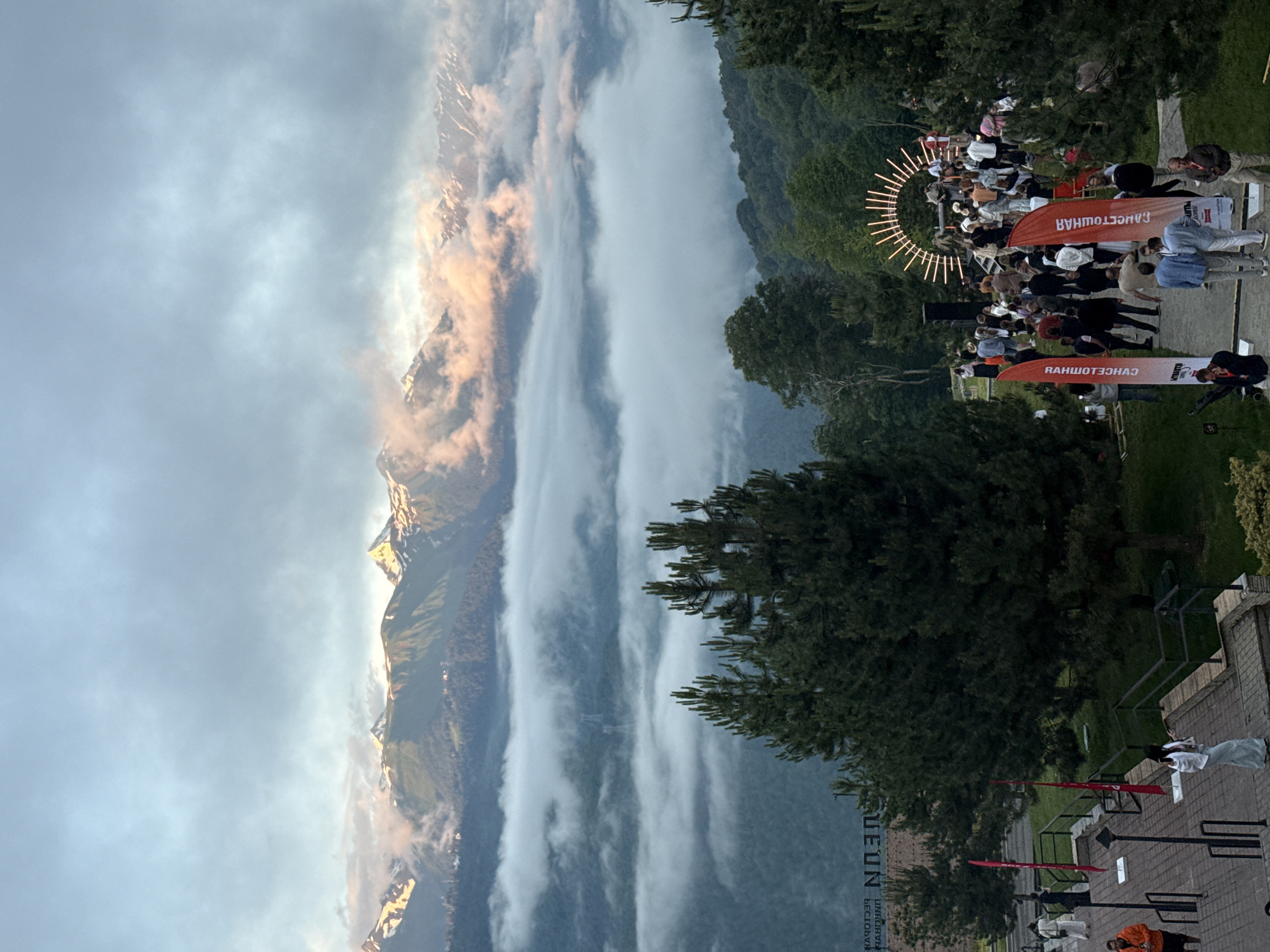 This is a photograph of an outdoor event in Estosadok, Russia. The scene is set against a dramatic backdrop of a mountain range partially obscured by clouds, with the peaks catching the warm light of what appears to be either sunrise or sunset. Lush green trees frame the right side of the image, suggesting a mountainous, forested environment.
In the foreground, a crowd of people are gathered. They are standing and mingling, with some facing a circular structure adorned with lights. Two large, orange banners are prominently displayed, one in the mid-ground and another further back. The banners have text printed on them, appearing to be in Cyrillic script and possibly reversed.
The overall atmosphere seems to be one of a gathering or festival, possibly an evening event given the lighting structure. The weather appears to be slightly overcast, contributing to the atmospheric lighting on the mountains. The people are dressed casually, and their body language suggests a relaxed social setting. The presence of a paved walkway and railings indicates a developed area within the natural landscape.