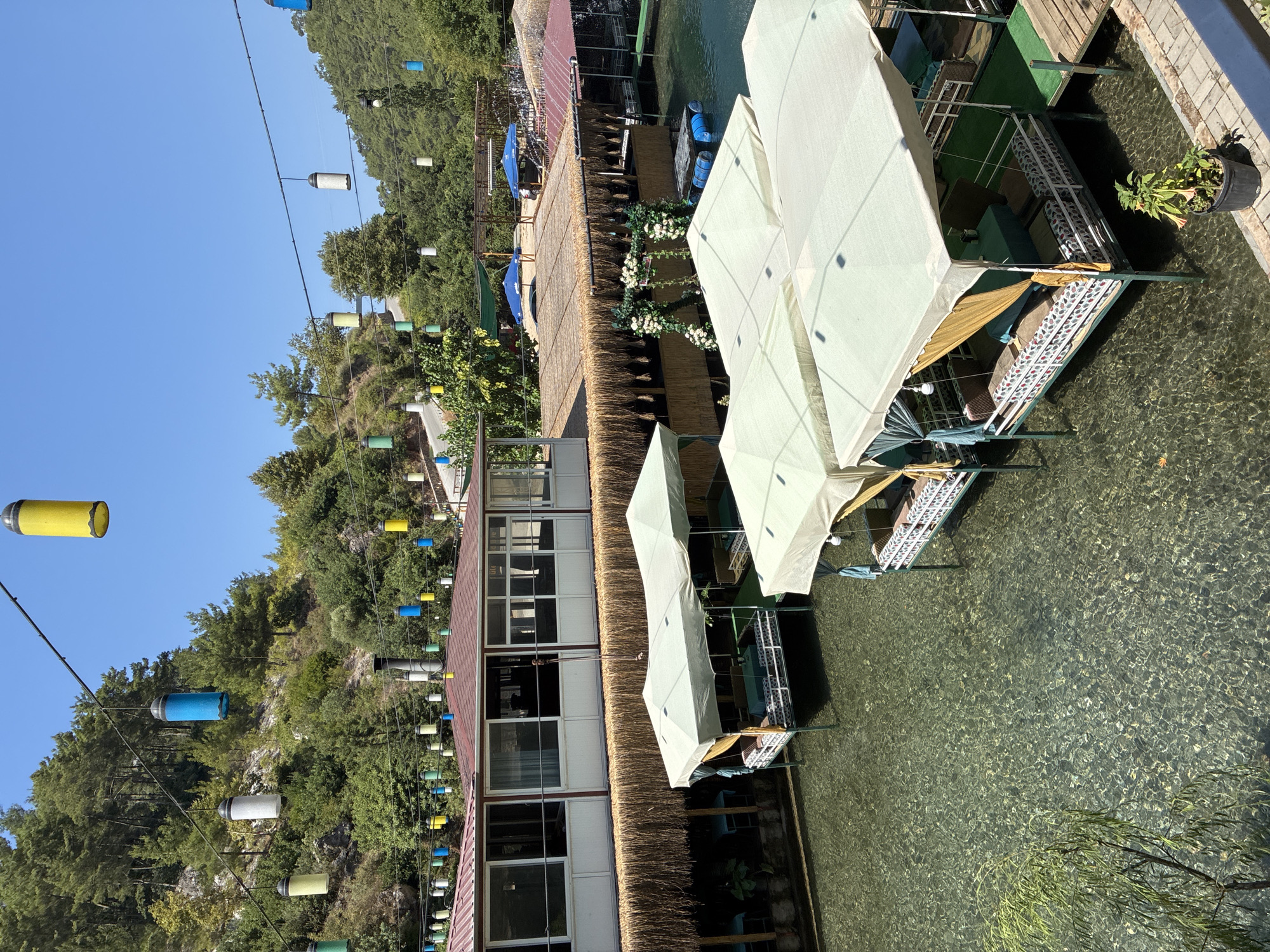 This is a photograph of an outdoor dining area situated on a body of clear water, likely a river or stream, in Alanya, Turkey. The setting appears to be a restaurant with several open-air dining platforms, each covered by a light-colored canopy. These platforms are elevated and suspended over the water, with seating arrangements visible on them. In the background, there is a building with a red roof and large windows, as well as lush green hills covered in trees. String lights, in various colors such as yellow, blue, and white, are visible hanging overhead, suggesting it might be an evening or a festive atmosphere. The water is transparent, allowing the rocky bottom to be seen. A small tree with green leaves is present in the foreground. The overall impression is one of a serene and picturesque dining experience in a natural environment.