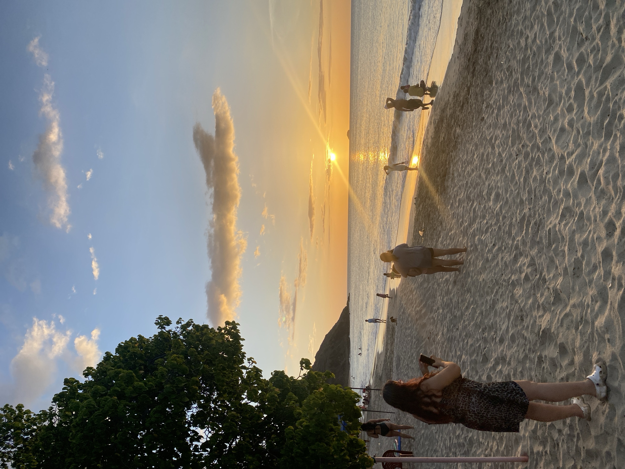 The image depicts a beach scene at sunset. The sun is low on the horizon, casting a warm, golden glow across the sky and water. The sky is a gradient of blue and orange, with scattered clouds, one of which is particularly prominent and elongated. The ocean meets the sandy shore with gentle waves.
Several people are visible on the beach. In the foreground, a woman with long, dark hair is standing and looking at her phone, wearing a patterned dress and white Crocs. Further up the beach, a man and woman are walking or standing, their silhouettes visible against the sunset. Other individuals and groups are scattered along the shoreline, some appearing to be wading in the water. A large, lush green tree dominates the left side of the frame, its leaves catching the sunlight. A dark, mountainous landmass is visible in the distance on the right side of the image, partially silhouetted against the sky.
The overall mood is tranquil and serene, characteristic of a sunset at a tropical location. The time of day is clearly sunset, and the weather appears to be clear with some cloud cover. The location cues suggest a coastal area, with the presence of the sea, sand, and palm-like trees. There is no visible text in the image.