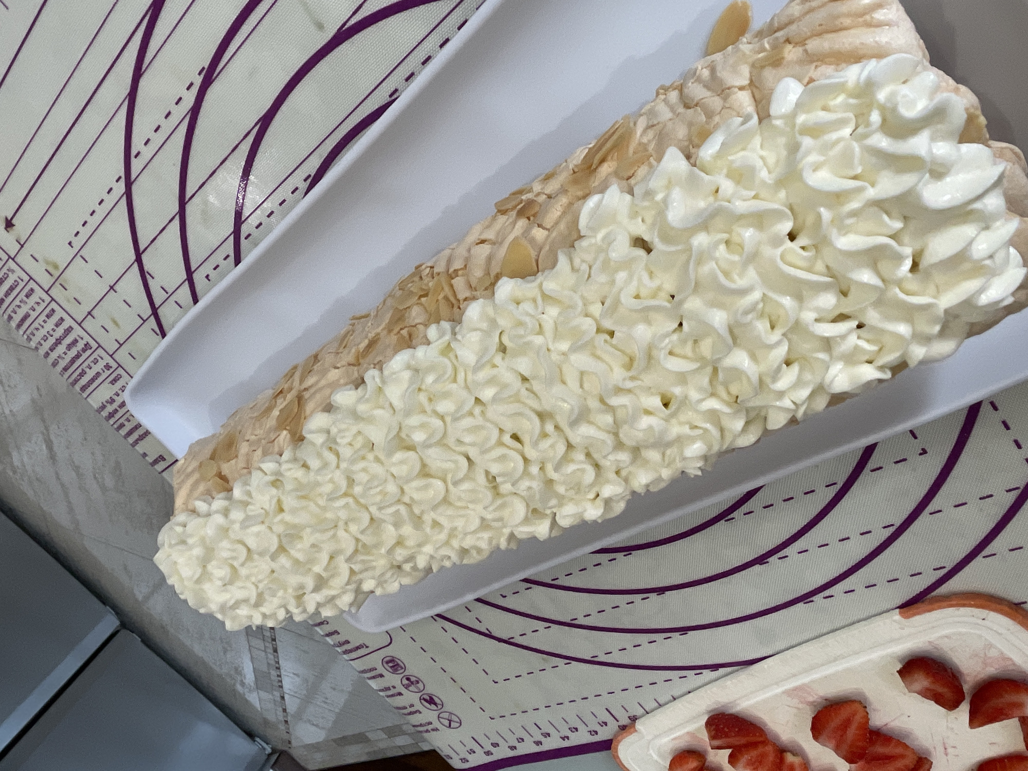 An overhead shot shows a meringue roulade covered in piped whipped cream and flaked almonds, resting on a white platter. To the left, a white tray holds sliced strawberries. The roulade and platter sit on a purple and white baking mat with circular markings and measurement lines. In the background, a grey refrigerator and a tiled countertop are visible. The scene appears to be in a kitchen in Saint Petersburg, Russia.