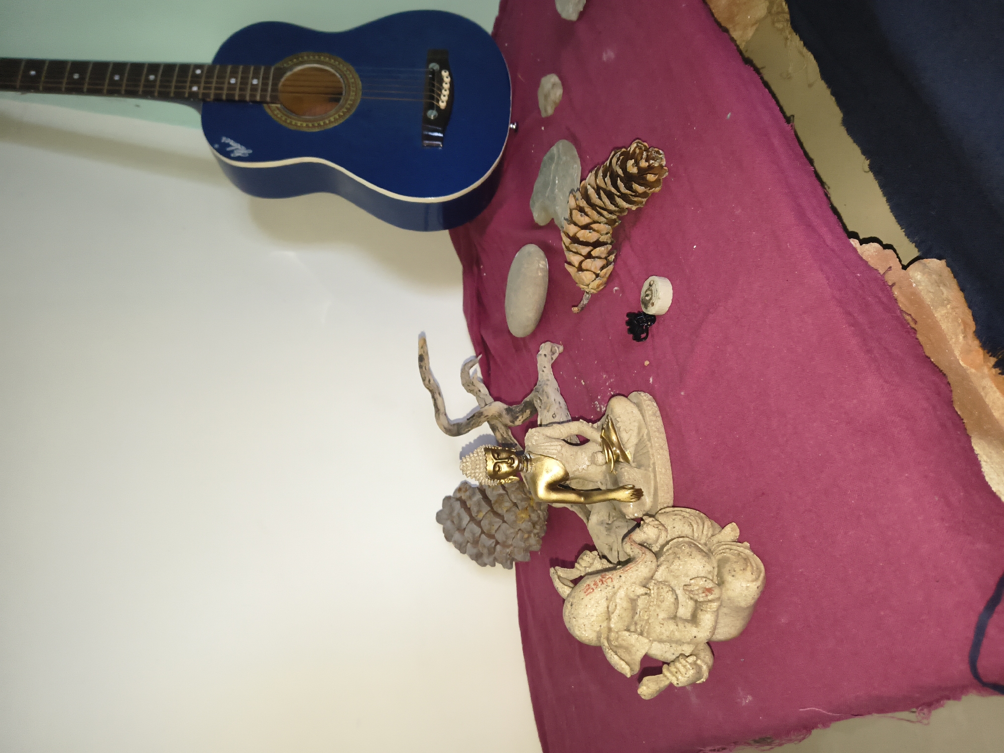 The image depicts an indoor scene with a collection of objects arranged on a magenta cloth. A blue acoustic guitar leans against a wall in the background, to the right. In the foreground, on the cloth, are two statues: one of Ganesha on the left, and a seated Buddha in the center. To the right of the Buddha statue, there are several natural elements, including a pine cone, stones, and a piece of driftwood. The background wall is plain and white, with a hint of mint green on the far right. The overall impression is one of a decorative or spiritual arrangement. No specific location in the city of Haini, India is identifiable from the image.