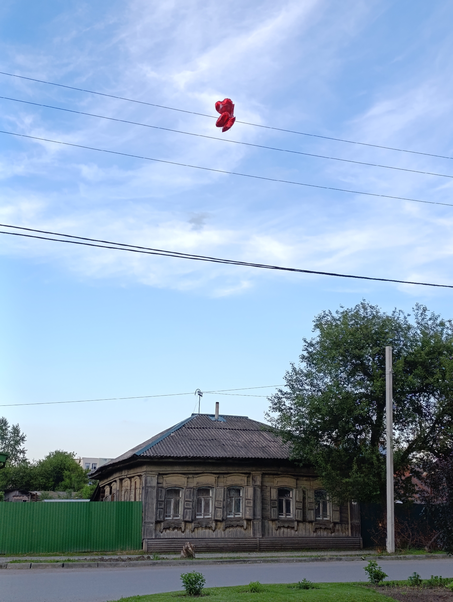 The image presents a quiet street scene in Tara, Russia, under a bright blue sky adorned with scattered white clouds. High in the frame, a cluster of bright red balloons is visibly snagged and tangled across several dark overhead power lines, contrasting sharply with the sky.
Below, the main subject is an old, traditional wooden house, typical of Russian architecture (an izba). Its weathered wooden siding appears unpainted, and it features intricate, decorative carvings around the windows, which are fitted with closed wooden shutters. The roof is dark and textured, likely made of corrugated material or slate tiles, and a small antenna is visible on top.
To the left of the house, a vibrant green corrugated metal fence marks the property line, behind which smaller trees are visible. A large, leafy green tree stands to the right of the house, partially obscuring it, and a tall concrete utility pole is positioned next to the tree. In the foreground, a paved road stretches horizontally, flanked by a narrow strip of green grass with a small bush. The soft, diffuse lighting suggests the time of day is either late afternoon or early evening. There are no people visible, and no discernible text appears in the scene.