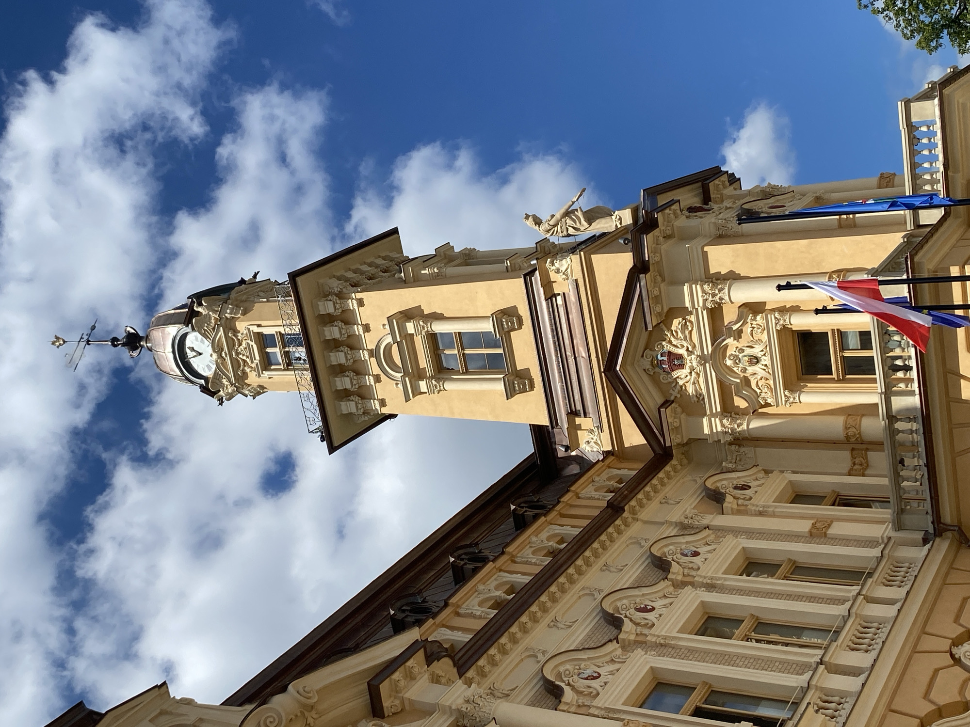 The image shows the Nowy Sącz City Hall, a historic building located in Nowy Sącz, Poland. The facade is predominantly yellow with ornate white stucco decorations. A prominent clock tower with a copper dome rises from the building, topped by a weather vane. The tower features decorative sculptures and windows. Below the tower, there is a balcony with a wrought-iron railing. Several flags are visible, including the Polish flag. The building is set against a bright blue sky with scattered white clouds. The architecture appears to be in the Secessionist style, also known as Art Nouveau. The overall impression is of a grand, well-maintained historical landmark on a clear day.