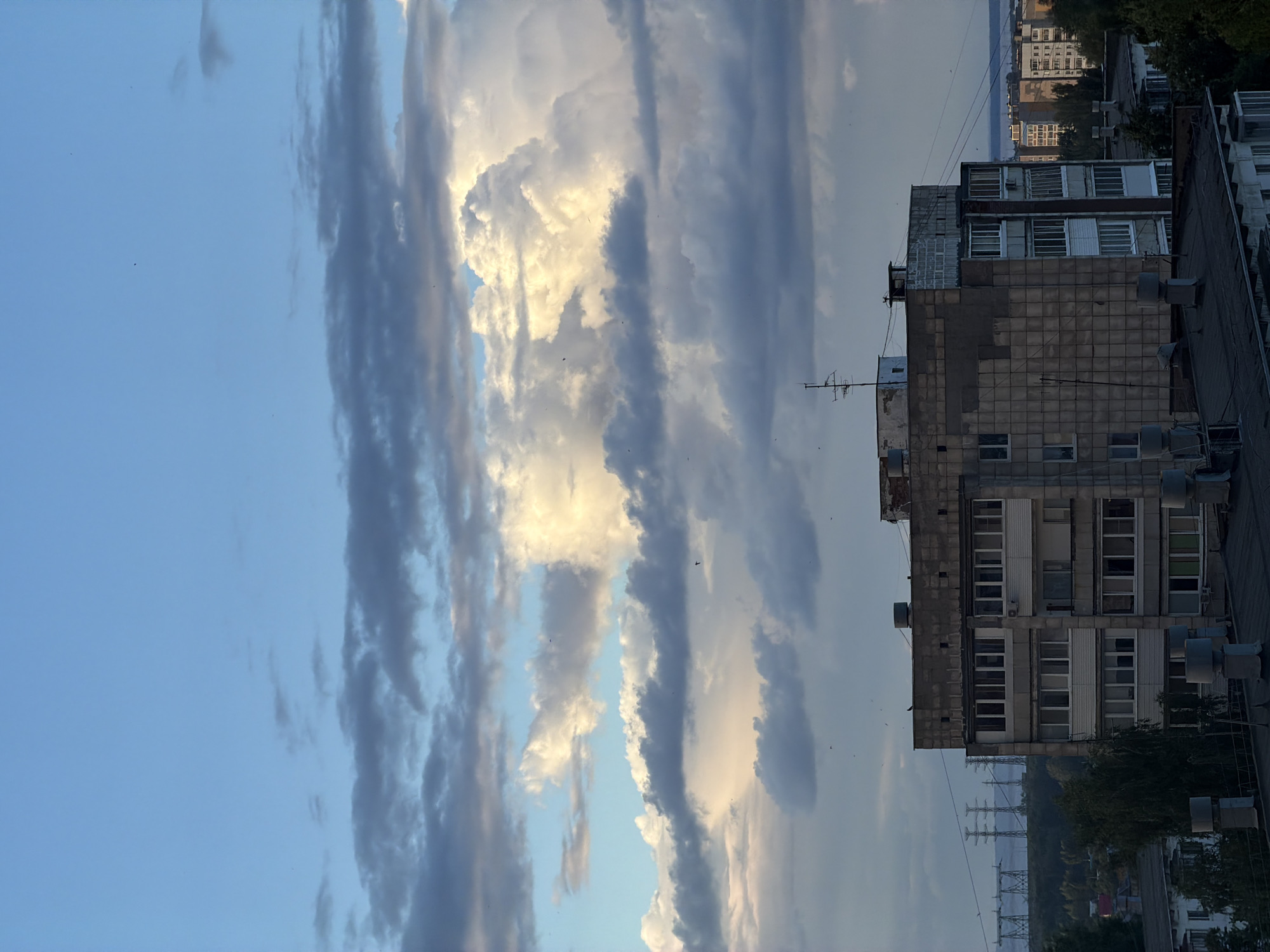 This media file captures a view of buildings and a cloudy sky in Chaikovsky, Russia. The orientation of the image is rotated 90 degrees clockwise.
The subjects are primarily buildings, appearing to be apartment blocks, with varying architectural styles and conditions. Some buildings have a rough concrete facade, while others feature more uniform window arrangements. Visible objects include what appear to be air conditioning units attached to the exteriors of the buildings, and an antenna on one of the rooftops. In the background, more distant buildings and a body of water are visible, possibly a river or reservoir, along with a line of power pylons. The sky is filled with dramatic, elongated clouds, some illuminated by sunlight, creating a contrast between bright whites and dark grays. A few birds are discernible as small dark specks against the sky.
The time of day appears to be late afternoon or early evening, given the quality of light illuminating the clouds. The weather is mixed, with sunshine breaking through a cloudy sky. No people are visible in the frame. The scene evokes a sense of urban environment under a dynamic sky, potentially a quiet moment before dusk. There is no visible text in the image.