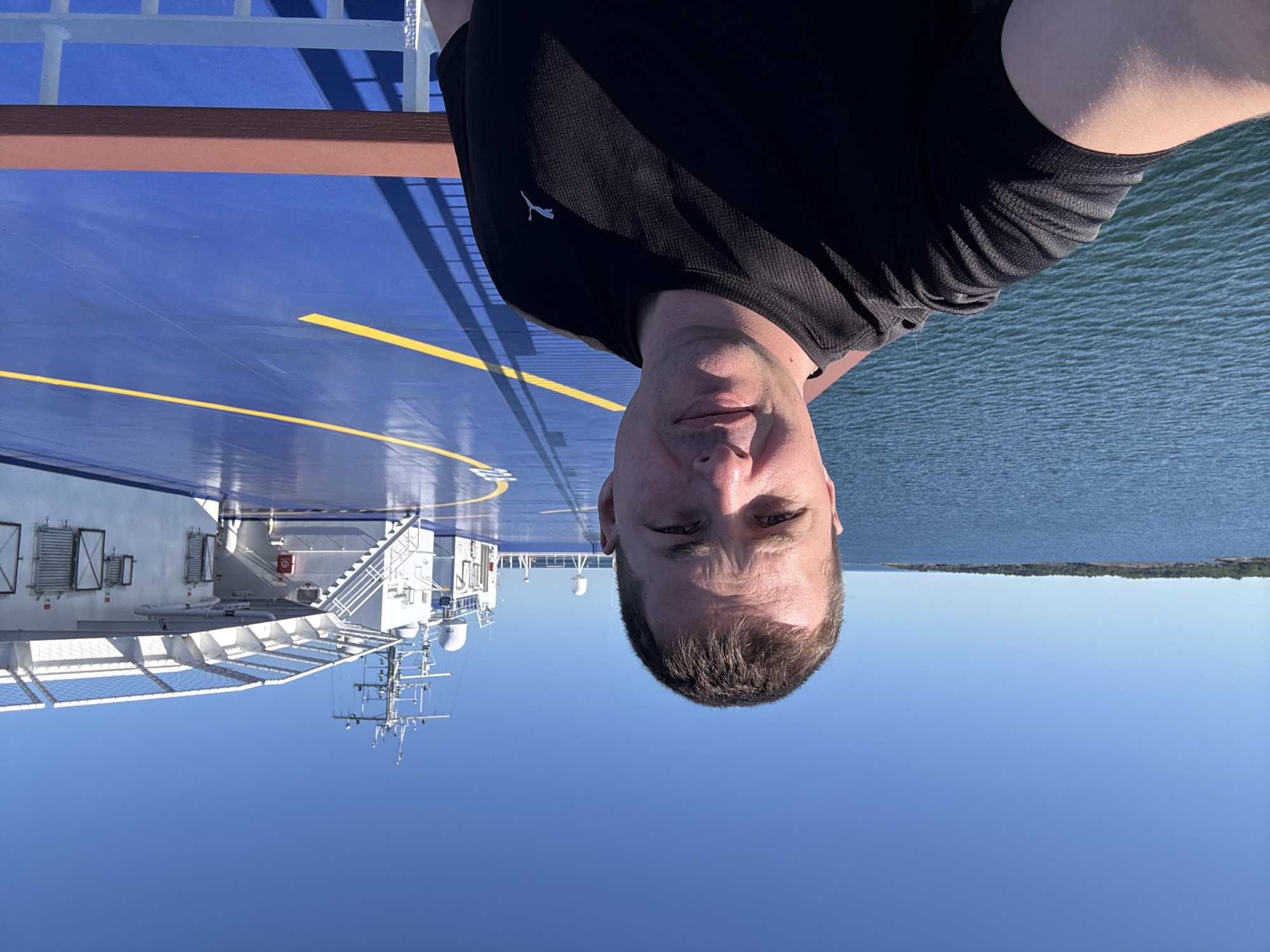 A man is taking a selfie on the deck of a ferry. He is wearing a black t-shirt and is smiling at the camera. The ferry deck is painted blue with yellow markings. In the background, there is the ocean, a distant island, and the upper structures of the ferry, including a helideck and a navigation tower. The sky is clear and blue. The scene appears to be in the waters near Karlskrona, Sweden, given the presence of ferries and the archipelago typical of the region.