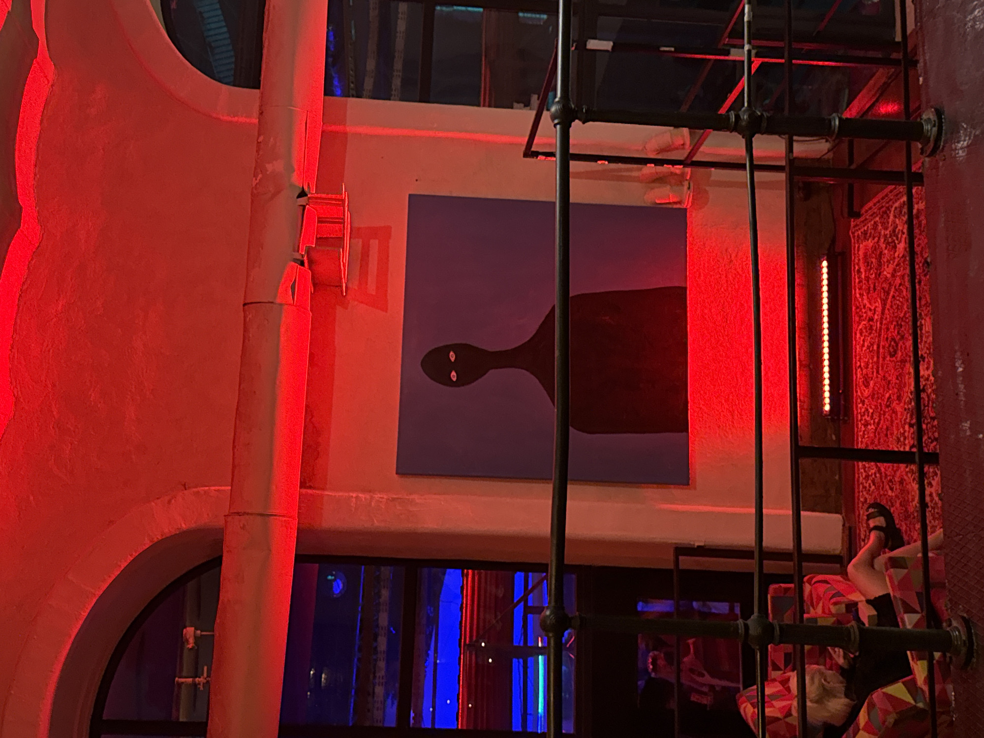 The image depicts an interior scene in Moscow, Russia, dominated by intense red ambient lighting. A large, rectangular painting hangs prominently on a light-colored wall, featuring a dark, silhouetted humanoid figure with an elongated neck and two distinct, glowing white-orange eyes, set against a pale purple background. Above the painting, industrial-style piping runs horizontally across the upper wall, beneath which linear red lights are installed.
To the left of the painting, a set of arched windows reveals an exterior or adjacent space illuminated by blue light, with reflections of the red-lit interior visible on the glass. Further down, behind a black metal railing, a blonde individual is seated in a multi-colored armchair, covered in a geometric pattern of red, orange, yellow, blue, and black. The person wears dark clothing, possibly shorts, and black sandals, and appears to be looking down at something in their lap. On the floor in front of the seated person, a red, ornately patterned rug is visible. More linear red lights are positioned at floor level to the right, contributing to the pervasive red glow of the environment. The foreground features a dark, textured floor, and a black metal railing with decorative finials spans across the lower half of the image, indicating a mezzanine or elevated vantage point.