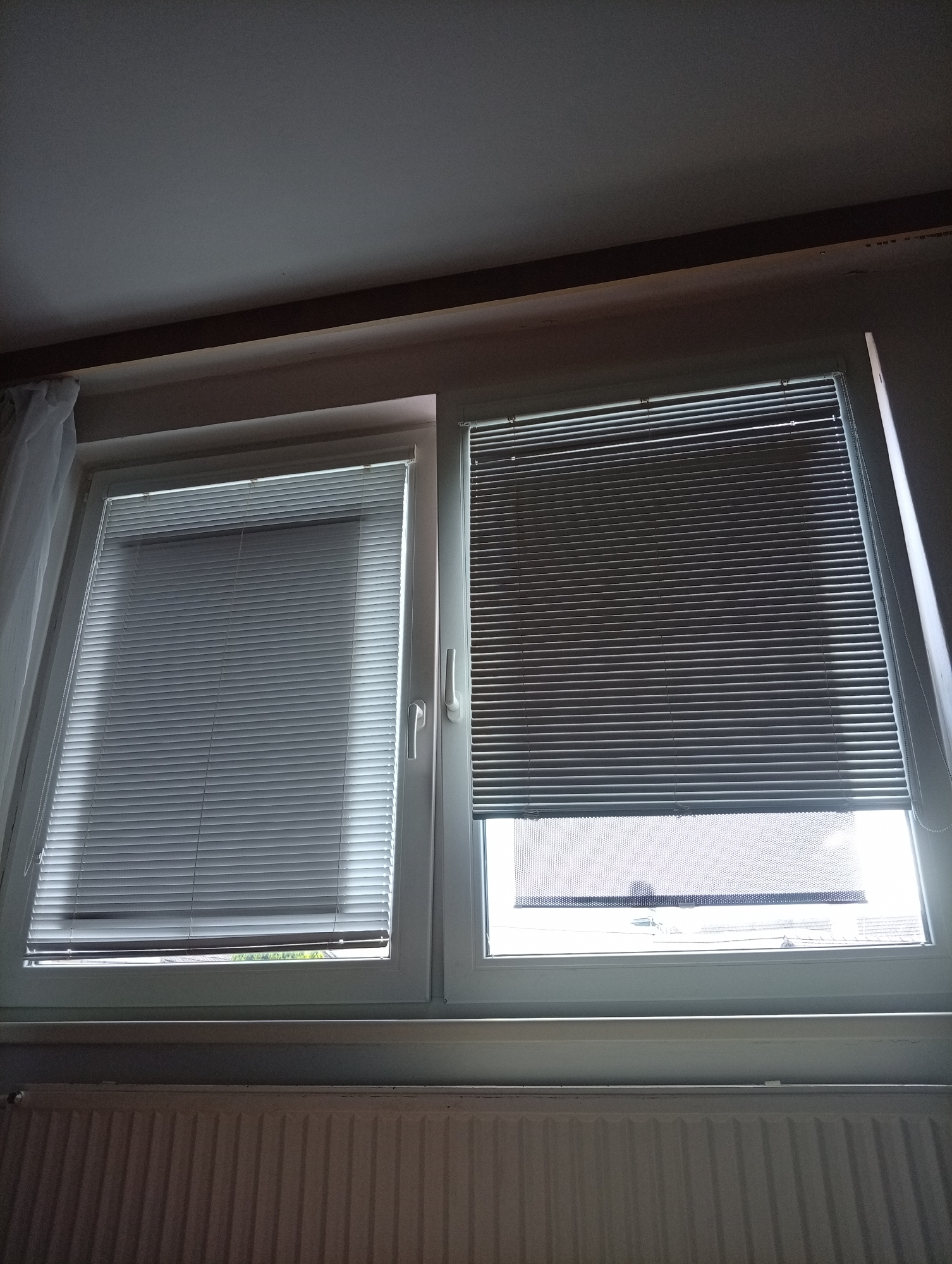 The image shows two closed blinds on a window in a room. The left blind is a light color, while the right blind is a darker shade. The window is part of a modern white frame, and to the left, a sheer white curtain is visible. Below the window is a white radiator with vertical fins. The overall lighting suggests an indoor environment, likely a residential setting. The scene does not contain any people or overt actions. The provided context places this scene in Tišnov, Czechia, though this information is not visually discernible from the image itself.