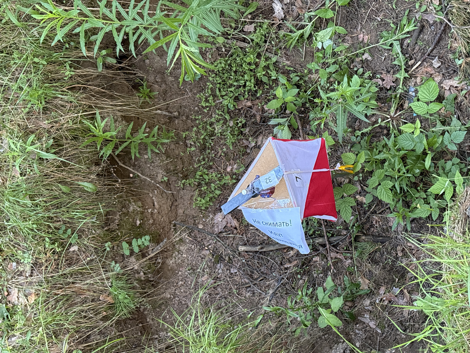 The image shows an orienteering marker in a natural environment, possibly a forest or park. The marker is a triangular prism with white and red sides, and a corkboard top where a punch is attached. A white flag with Russian text is also attached to the marker. The ground around the marker is covered with dirt, leaves, and grass, and there is a steep embankment with dense vegetation in the background. There are no people visible in the image. The text on the flag is "Не снимать! Клуп Я Неизвестных", which translates to "Do not remove! Club Unknowns". This suggests the marker is part of a competition or event organized by a club. The location is likely in the city of Chaikovsky, Russia, as indicated by the text on the flag.