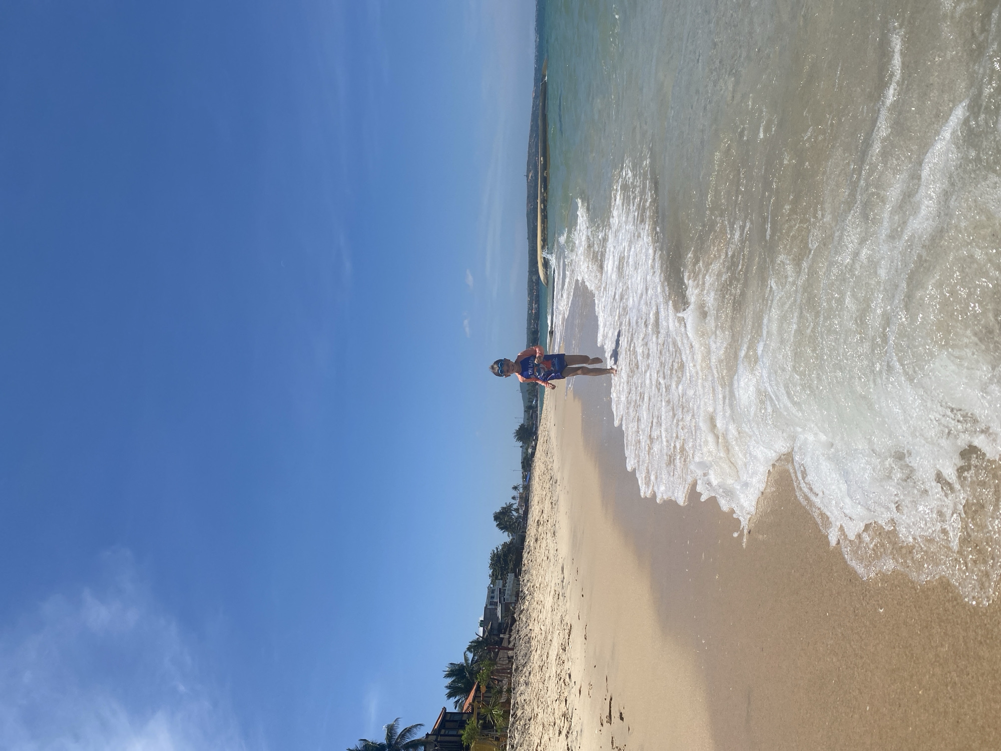 A young child, wearing a rash guard and swim trunks, runs along the shoreline on a sunny day at Phu Thuy Ward, Vietnam. The child is moving from right to left, facing the camera, with a bright blue sky and scattered clouds overhead. The beach is sandy, with a gentle wave washing ashore, creating foam and ripples. In the background, there are palm trees and buildings lining the coast, suggesting a developed area. The water is clear and turquoise, with a slight chop. The overall scene depicts a carefree moment of a child enjoying a day at the beach.