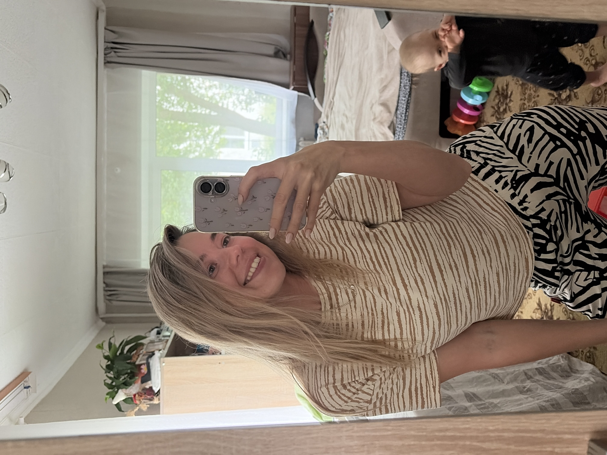 An indoor mirror selfie captures a smiling woman and a baby in what appears to be a residential room. The image is oriented sideways, with the woman's head to the left. The woman has long, light brown hair and is smiling brightly at the camera, which she holds in her left hand. She is wearing a cream-colored top with vertical brown stripes and patterned pants featuring a black and white zebra print. Her smartphone has a light-colored case adorned with a delicate floral pattern.
Reflected in the upper right portion of the mirror is a baby with light blonde hair, dressed in a dark, long-sleeved outfit. The baby is positioned on a patterned rug, looking towards the left, and a colorful stacking ring toy (red, orange, yellow, green, and blue rings) is visible on the floor next to them.
The background reveals a bright, sunlit room, likely a bedroom or living area. A large window with white frames shows lush green foliage outside, suggesting trees and an outdoor view. Gray pleated curtains or blinds are drawn near the top of the window. A bed with light-colored bedding (beige or cream) is partially reflected behind the baby. To the lower left, a wooden dresser or cabinet is visible, topped with a vibrant green potted plant. The overall atmosphere is cheerful and domestic. No discernible text is visible in the image.