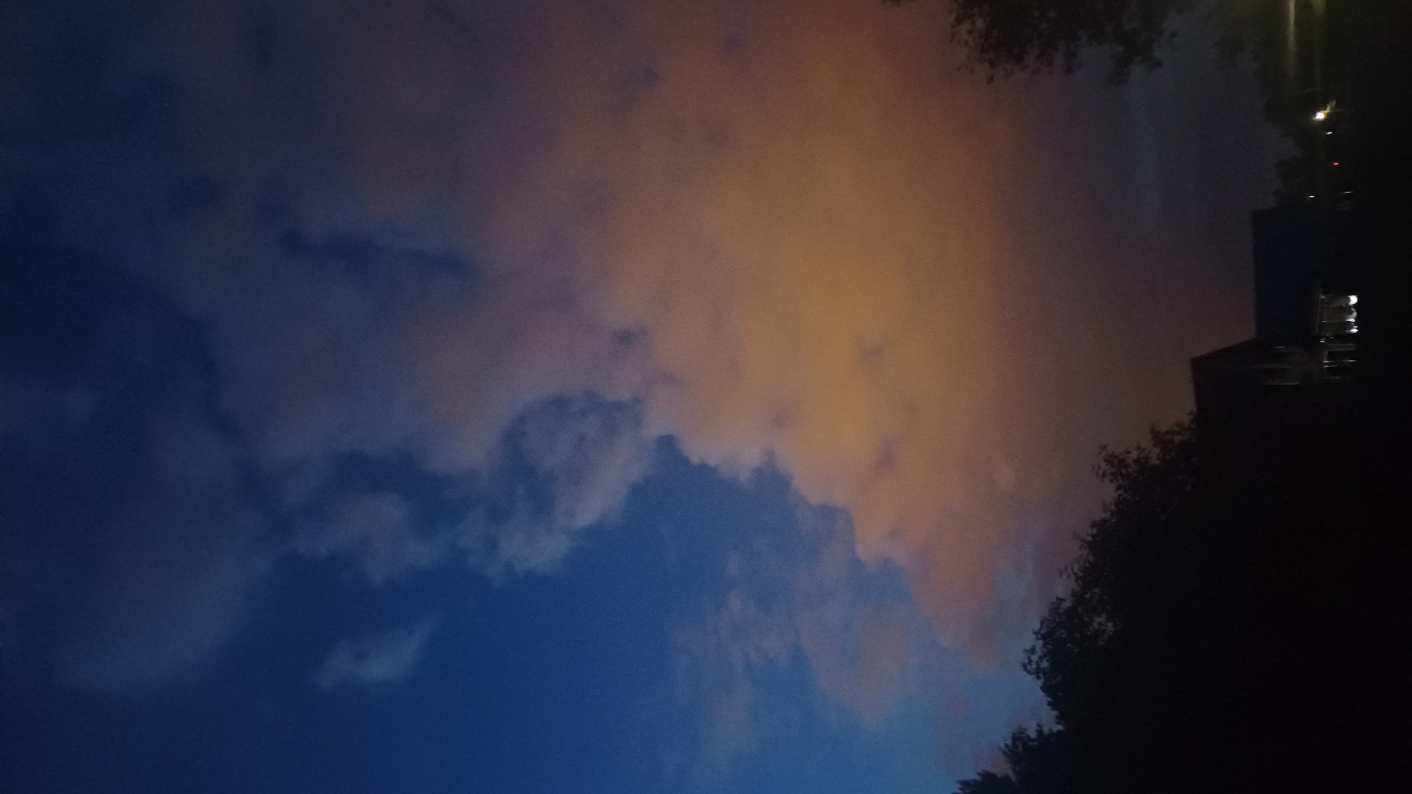 The image captures a dramatic sky scene at twilight or night, presented from a perspective rotated 90 degrees counter-clockwise. The lower portion of the image displays a deep, dark blue sky, partially obscured by distinct blue-grey clouds with defined edges and varied textures. Moving upwards and towards the right, the cloud formations transition into a vibrant orange and amber glow, indicating strong illumination from an unseen light source. This illuminated cloud layer creates a striking contrast with the cooler, darker blues of the lower sky.
On the right side of the image, silhouetted against the dark sky and glowing clouds, are dense tree canopies and what appears to be a building or urban structure. Several artificial lights are visible within the building's silhouette, notably a bright white light and smaller, less defined light sources, likely windows or streetlights. These artificial lights are the probable source of the orange glow reflecting off the clouds.
No human figures or specific activities are discernible; the scene focuses solely on the atmospheric conditions and minimal urban outlines. There is no visible text. The overall impression is one of a quiet, vividly illuminated night sky. The location context is Timokhovo, Russia.
