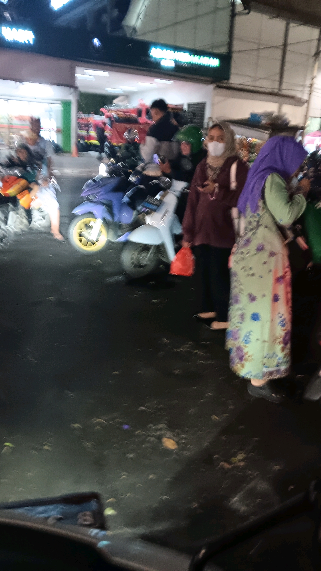 The image depicts a street scene at night in the city of South Jakarta, Indonesia, likely in front of a commercial establishment. Several individuals are visible, some of whom are wearing headscarves and face masks. One woman in the foreground wears a light green patterned dress and a purple headscarf. Another woman next to her wears a maroon long-sleeved top and a light-colored headscarf. Both women appear to be standing. Several motorcycles are parked in the scene, with one blue motorcycle prominently displayed. The ground is dark and appears to be asphalt or pavement, with scattered debris or litter. The background shows a building with illuminated signage, indicating a retail or service area. The lighting suggests it is nighttime, with artificial lights illuminating the scene. There is a sense of general activity, possibly indicating a marketplace or busy thoroughfare.