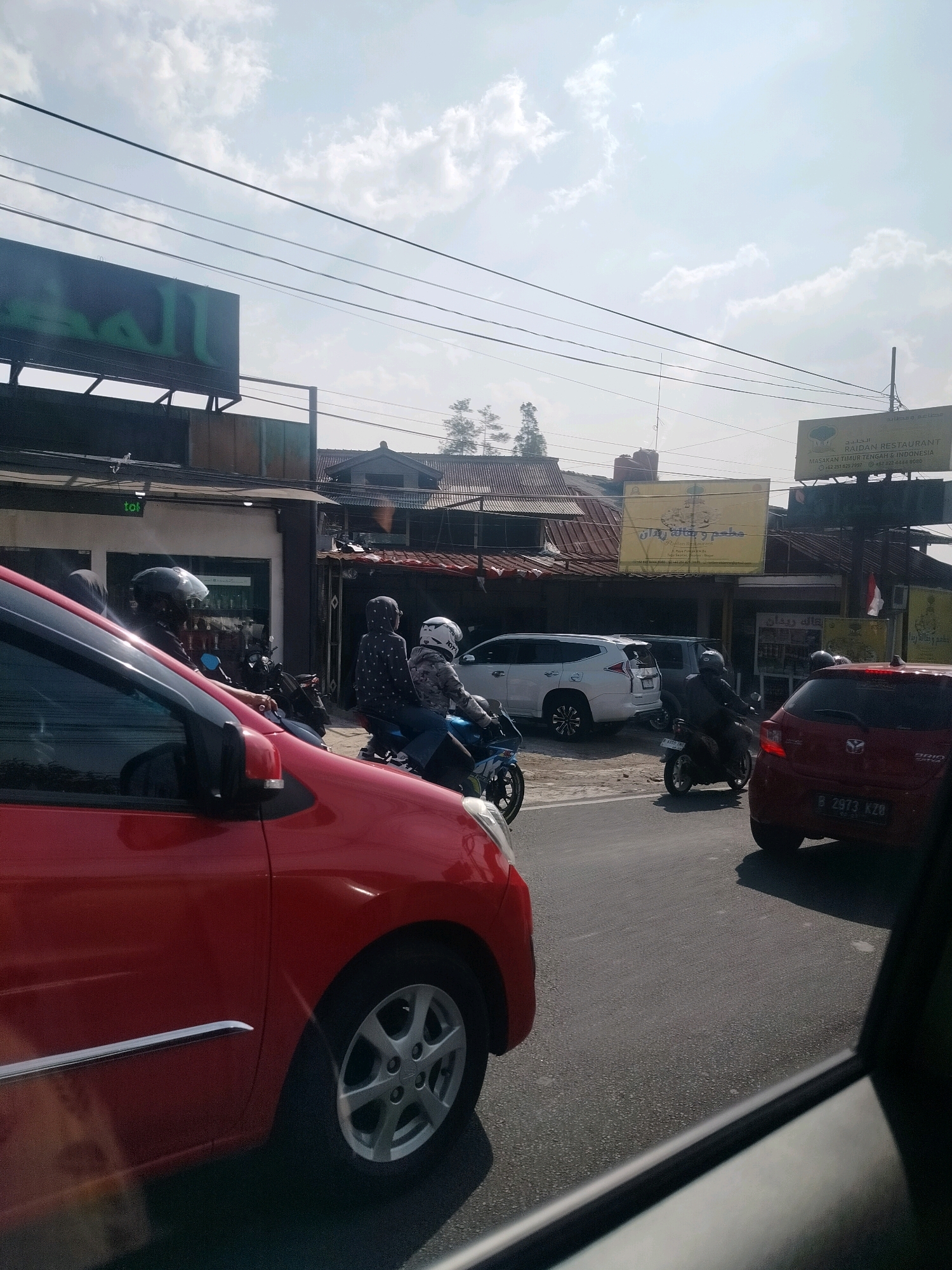 A street scene in Cisarua, Indonesia, on a sunny day with scattered clouds. In the foreground, the side of a red car is visible, showing its wheel and door handle. On the road, traffic is moving. Several motorcycles with riders are present. One rider wears a black helmet and a dark outfit. Another rider has a white helmet and a patterned jacket. A white SUV is parked on the side of the road, and a red car with a license plate reading "B 2973 KZG" is also in view. Overhead, power lines crisscross the sky. Buildings line the street, including a restaurant with signage that includes Arabic script and English text reading "RAIDAN RESTAURANT" and "MASAKAN TIMUR TENGAH & INDONESIA." Another sign with green Arabic lettering is visible on a building in the background. The overall environment appears to be an urban or semi-urban setting.