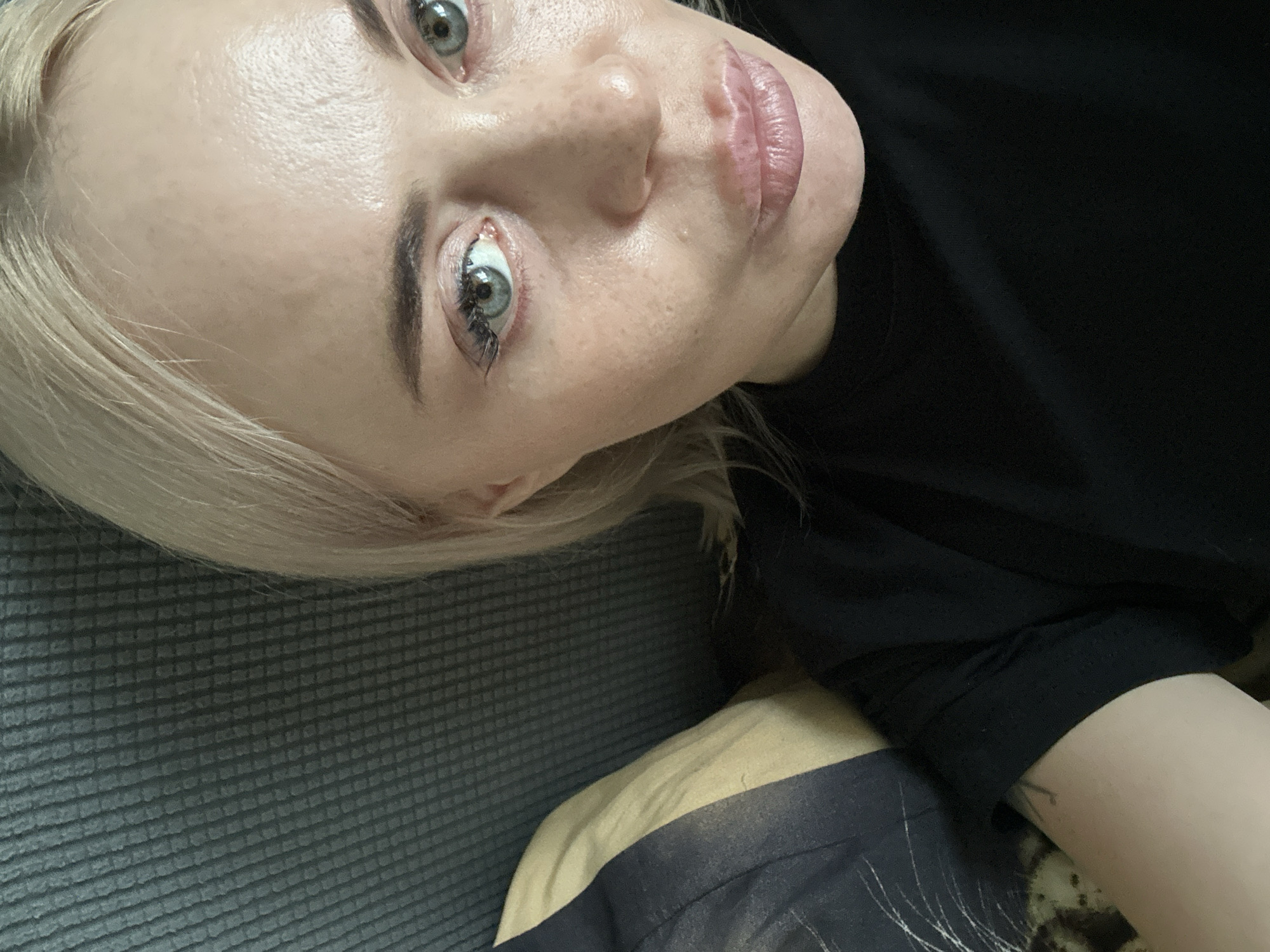 A close-up, overhead shot shows a pale-skinned woman with platinum blonde hair and light blue eyes. She is wearing a black t-shirt and is lying on a textured grey sofa with a striped pillow behind her. The lighting is soft, creating a gentle highlight on her forehead and nose. Her makeup is minimal, with a touch of mascara and a light pink lip color. The overall atmosphere is casual and relaxed.