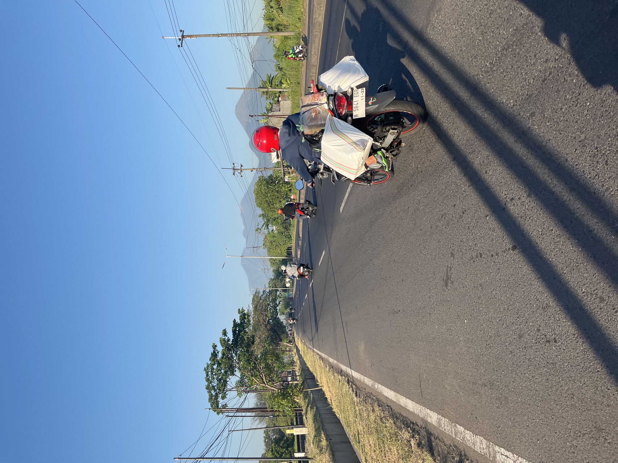 The image depicts a road in Siring, Indonesia, under a clear blue sky. The perspective is from the back of a motorcycle, looking ahead down a paved road. Several other motorcycles are visible, some carrying passengers and luggage. The road has a white line marking the lane edge. On the left side of the road, there is a grassy embankment with a concrete drainage channel, followed by trees and utility poles with numerous wires. In the distance, mountains are visible. The motorcycle in the foreground has a driver wearing a blue jacket and a red helmet, and is carrying two large white bags secured to the rear, along with other items. The license plate of this motorcycle is visible and reads "N 5841 EGB 11-29". The scene appears to be set during daylight hours, likely in the morning or late afternoon, given the long shadows cast by the utility poles.