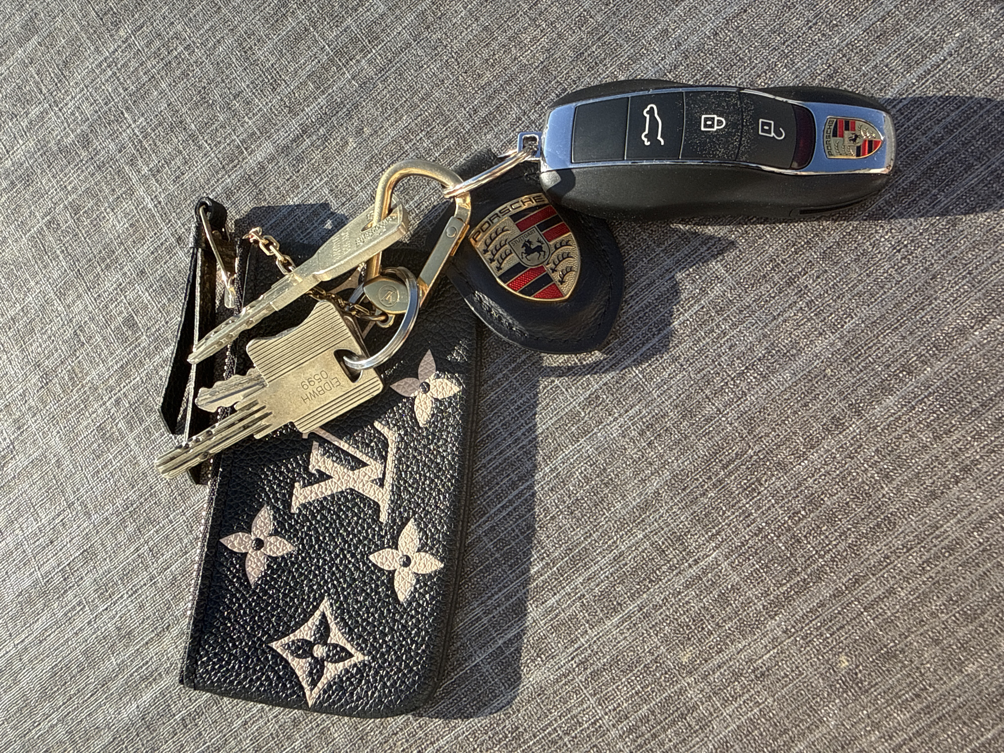 The image captures a set of luxury keys and a key case resting on a textured, light gray-brown woven fabric surface under bright, direct sunlight, casting distinct shadows. The scene is set in Käerjeng, Luxembourg.
The primary subjects include a black Porsche car key fob, a collection of metal keys on a gold-colored carabiner-style keyring, and a black Louis Vuitton key case. The Porsche car key fob features silver accents and visible buttons for unlocking, locking, opening the trunk, and a car-shaped symbol. A small Porsche crest, displaying "PORSCHE" and "STUTTGART," is integrated into the fob's design.
Attached to the keyring is a separate black leather keychain with a larger, detailed Porsche crest, also bearing the "PORSCHE" and "STUTTGART" text. Among the conventional metal keys, one is marked with "EIDBWH 0599," and another prominent key shows "6550 HAGOLA." A small gold charm with the "LV" monogram is also visible on the keyring.
The keys are positioned partially on a black Louis Vuitton key case, which features the brand's iconic monogram pattern in a tan or beige hue. This pattern includes the "LV" initials and characteristic quatrefoil and flower symbols. The overall impression is one of high-end accessories.