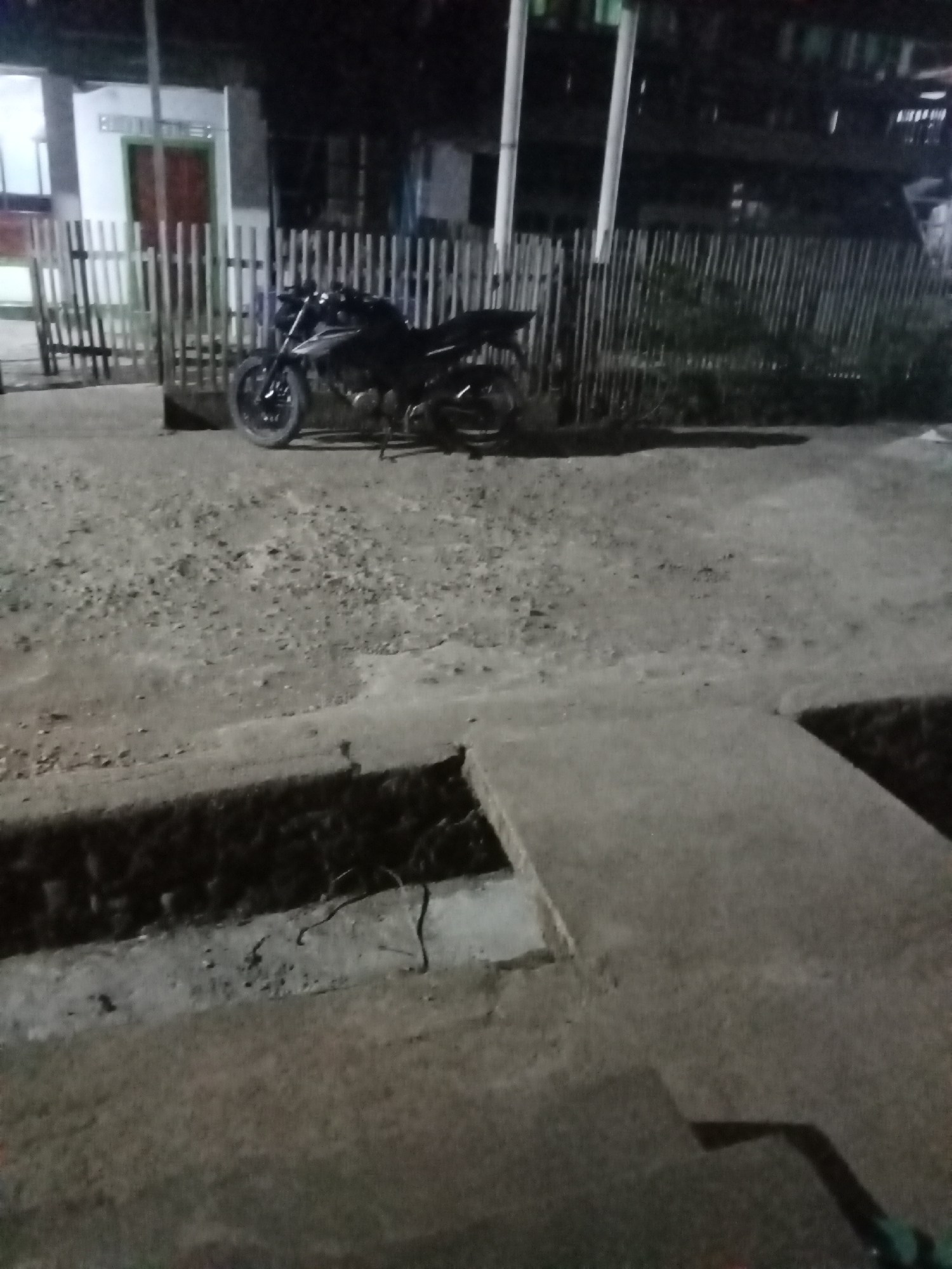 A black motorcycle is parked on a gravelly surface in front of a wooden fence. Behind the fence, a white building with red doors is visible. The scene appears to be an outdoor residential area at night, possibly in the city of Setia Jaya, Indonesia. The lighting is dim, casting shadows on the ground. There are no people visible in the image, and no actions or interactions are taking place. The environment is characterized by rough terrain and simple construction.