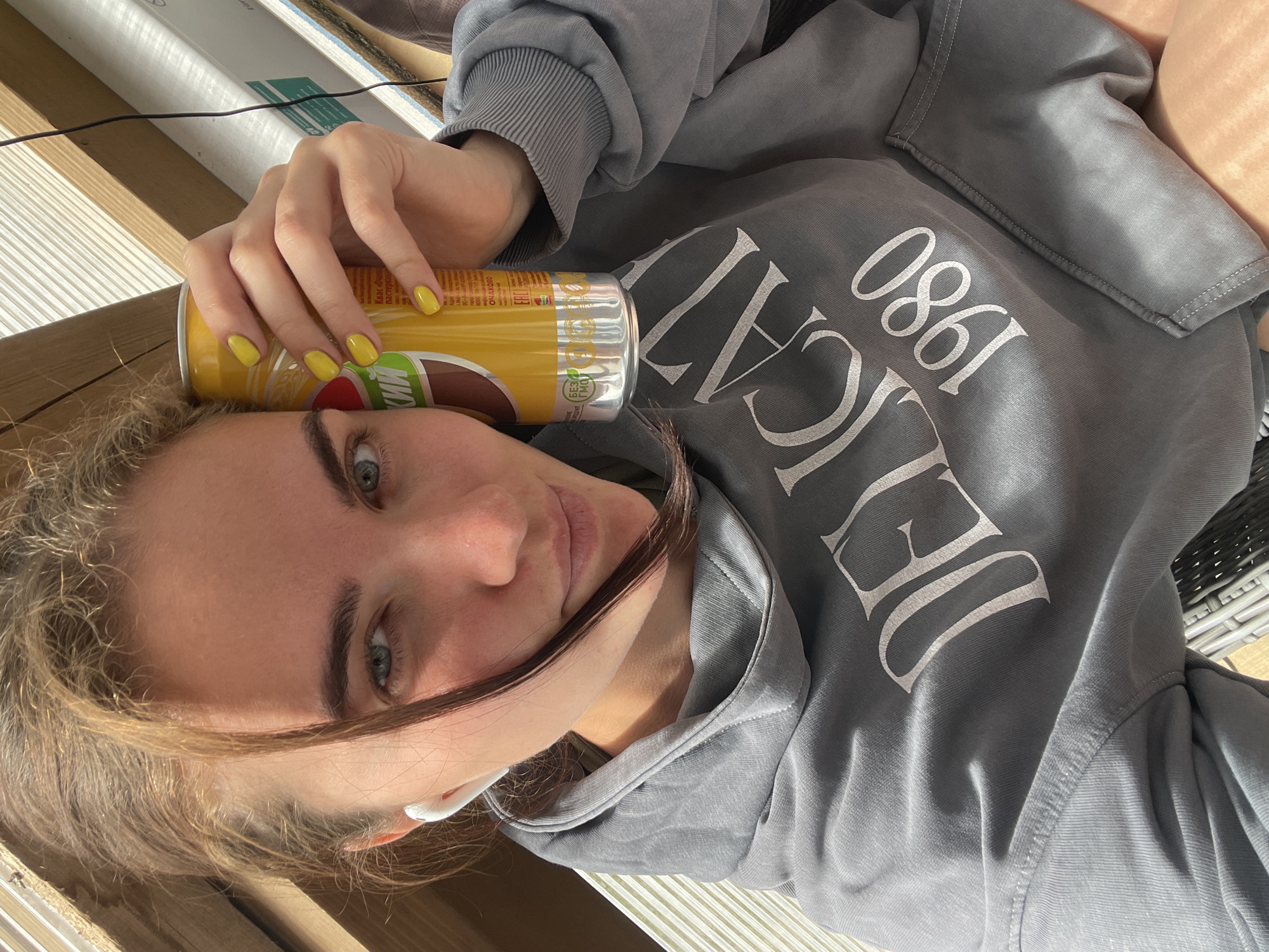 A young woman with blonde hair, blue eyes, and light skin is taking a selfie. She is wearing a grey hooded sweatshirt with the words "DELICATE 1980" printed in white on it. She is also wearing white earbuds. In her right hand, she is holding a yellow can of what appears to be a beverage. The background is blurred but suggests an outdoor setting with wooden structures and some sort of ribbed material. There are no identifiable city landmarks or specific locations within Мосметаллоконструкция Онт, Russia visible in the image.