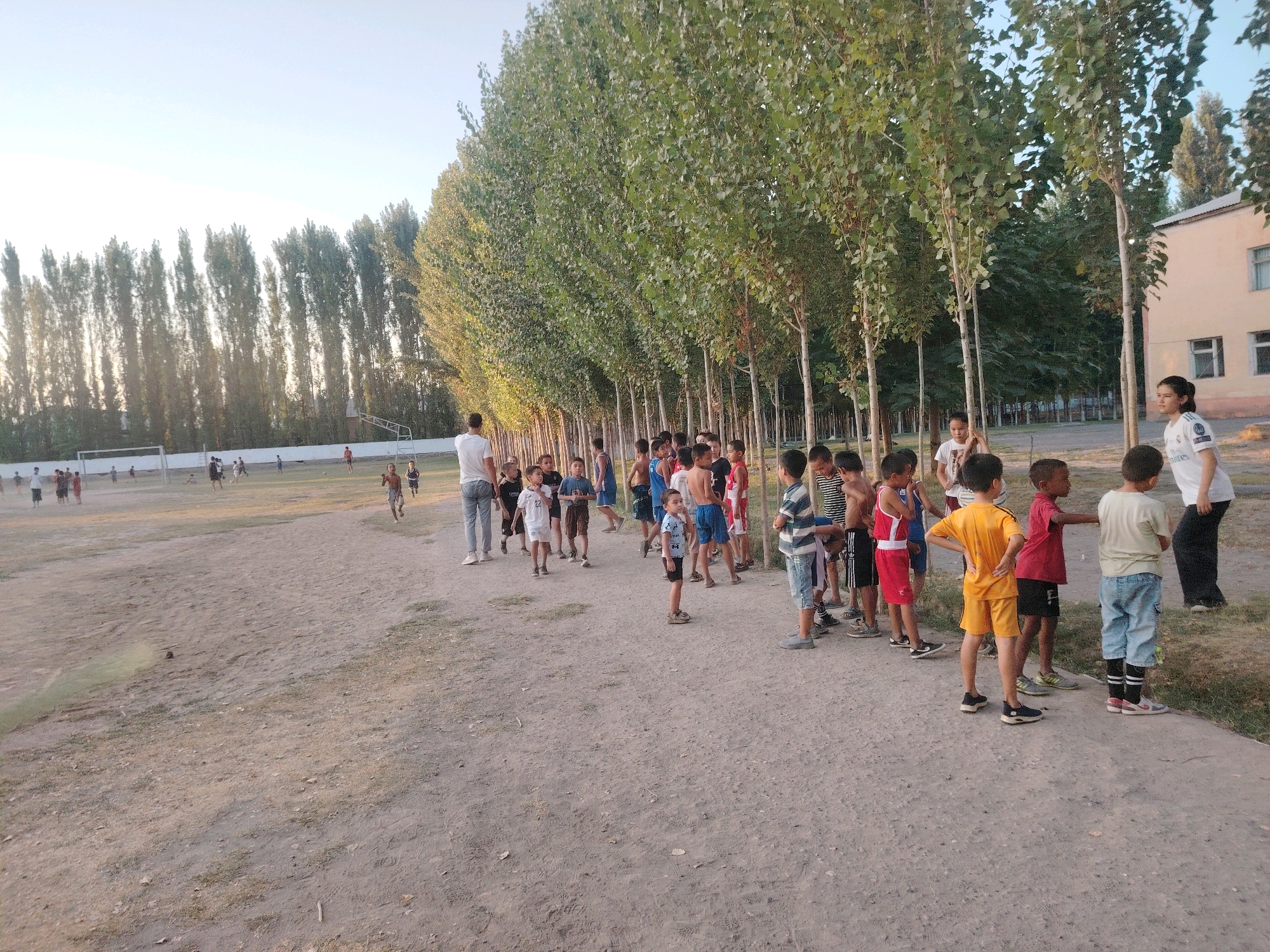 The image depicts an outdoor scene at a sports field in Paxtaobod, Uzbekistan. A group of approximately 20 children, mostly boys, are gathered near a line of tall trees. Some of the children are in athletic attire, such as shorts and t-shirts, and appear to be participating in or observing some activity. In the background, there are sports facilities including a goalpost and what seems to be a running track. A building, possibly a school or administrative facility, is visible to the right of the trees. The ground is dusty and appears to be unpaved. The lighting suggests it is either early morning or late afternoon.