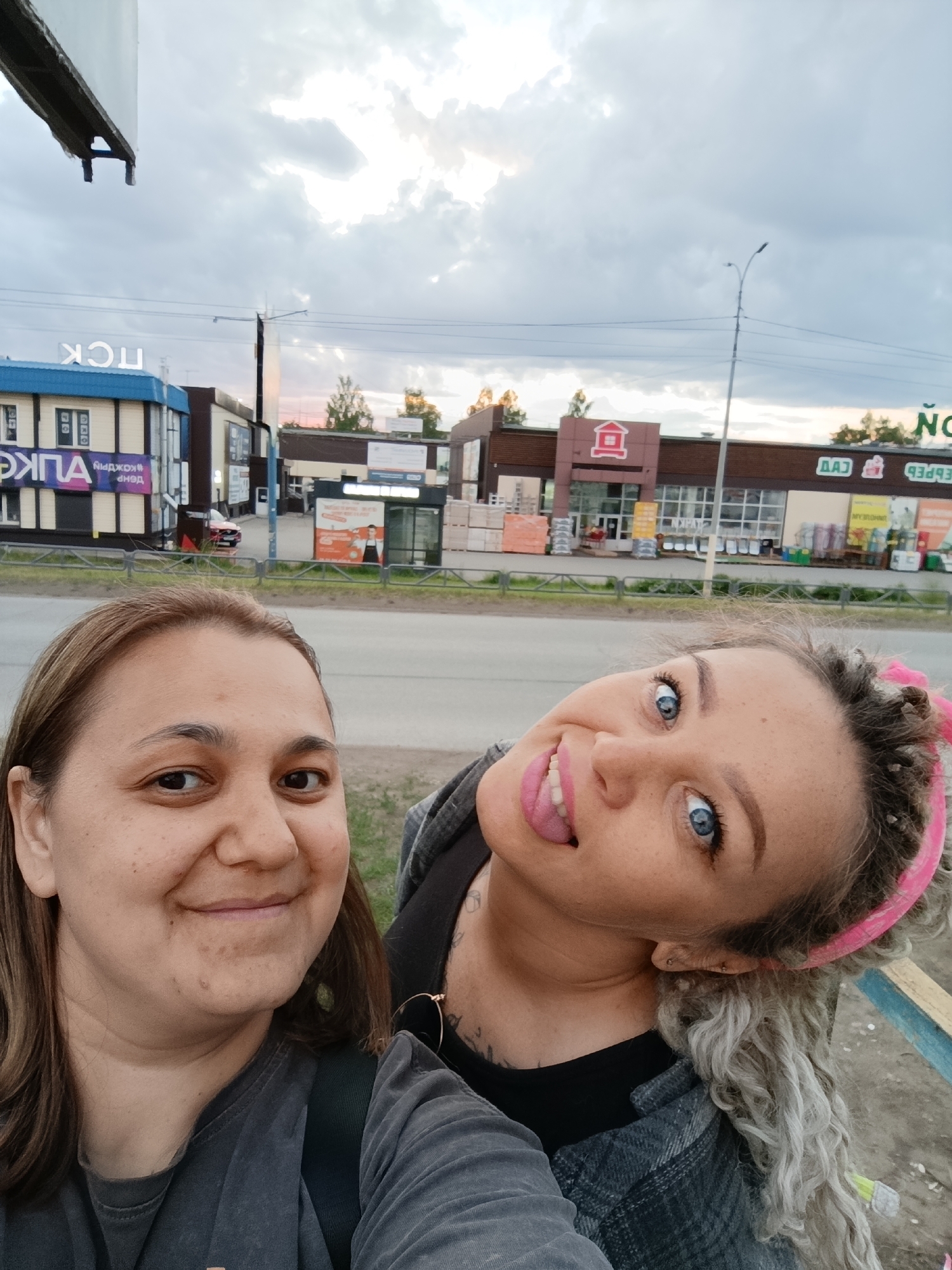 Two women take a selfie outdoors. The woman on the left has brown hair and a dark t-shirt. The woman on the right has long curly gray hair and a pink hairband. She is sticking her tongue out. Buildings are behind them. The first building on the left has blue signage, and other letters appear above the blue. The second building has a red logo. The third building has green signage. These buildings are across the street from where the women are standing. The location is likely a store in the city of Chaikovsky, Russia. A street light and a cloudy sky are above the buildings.