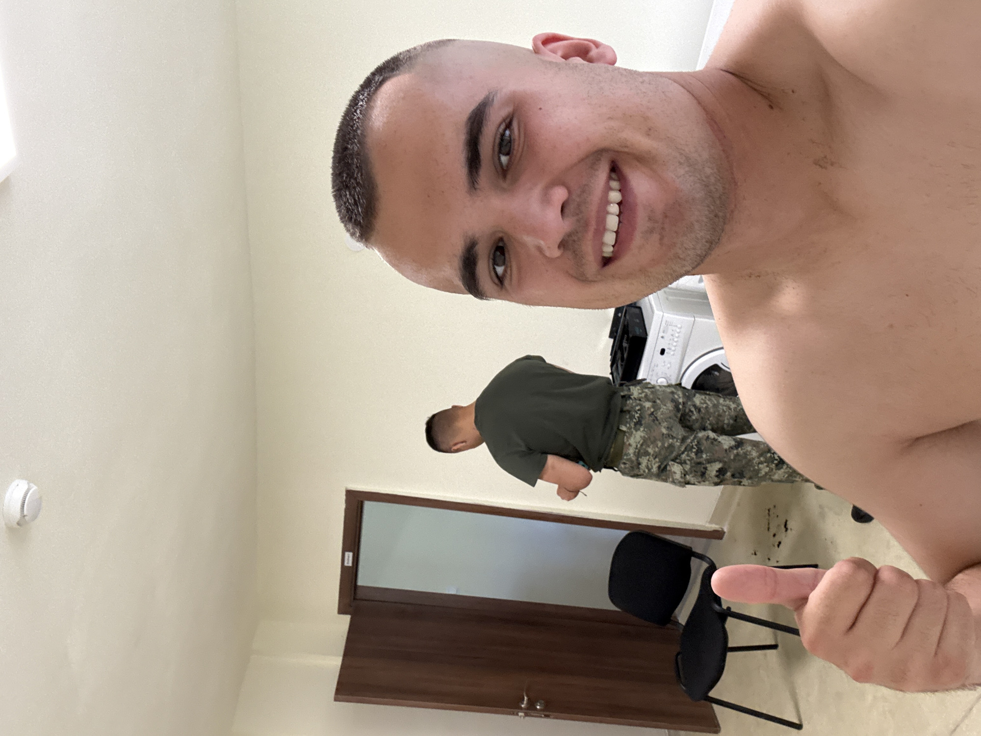 In an indoor room with pale yellow walls and a white ceiling with a smoke detector, a shirtless man with a buzz cut is giving a thumbs-up to the camera, smiling. He is positioned in the foreground, taking a selfie. In the background, another man wearing a green t-shirt and camouflage pants stands with his back to the camera, holding a pair of clippers and appearing to be cutting hair. To the left, a black chair with metal legs is placed near a dark brown door that is ajar, revealing another door inside. A white washing machine is visible behind the man with the clippers. The floor is tiled in a light color.