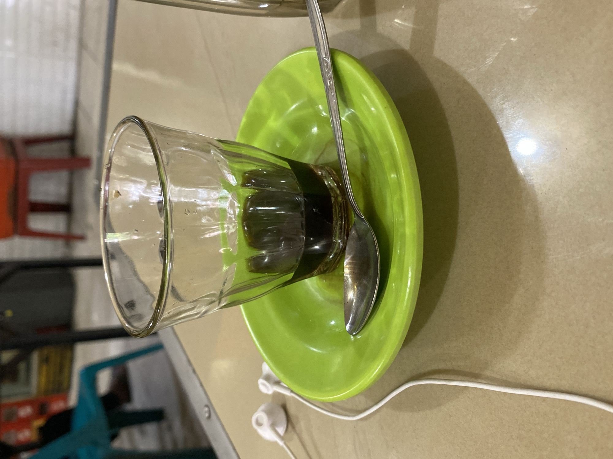 The image shows a close-up of a glass of dark liquid, likely coffee, on a bright green saucer. A silver spoon rests on the saucer next to the glass. To the left of the saucer, a pair of white earbuds with their white wire is visible. The objects are placed on a light-colored, speckled table. In the blurred background, several chairs, including a teal-colored chair, and tables suggest an indoor setting, possibly a cafe or restaurant. The overall lighting is dim, with a bright highlight reflecting off the table surface. The image appears to have been taken in Lhokseumawe, Indonesia, though no specific landmarks are identifiable. There are no visible people in the frame.