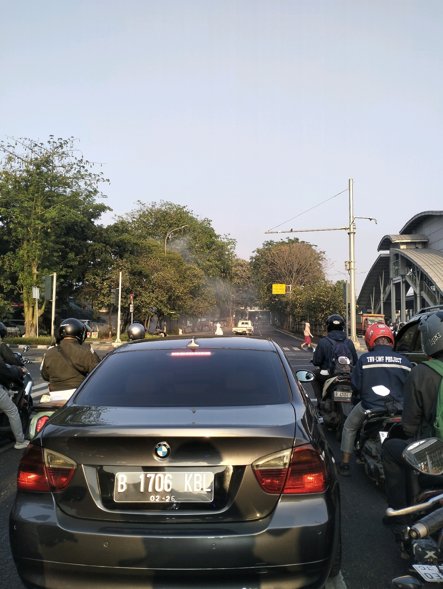 The image captures a traffic scene in East Jakarta, Indonesia, likely near a transportation hub given the elevated structure in the background. In the foreground, a dark-colored BMW sedan with license plate "B 1706 KBL" is stopped in traffic. Several motorcycles are also present, with riders wearing helmets. One rider on a motorcycle with license plate "B 4524 T" is wearing a dark blue jacket with reflective stripes and text that reads "TRY-LWF PROJECT". Another motorcycle rider is wearing a red helmet. Trees line the street, and a traffic light is visible. Smoke is rising from behind a tree on the right side of the image, obscuring part of the scene. The sky is clear and a bright, pale blue. An elevated walkway or bridge structure with a curved roof is visible on the right, suggesting proximity to a train station or similar infrastructure.