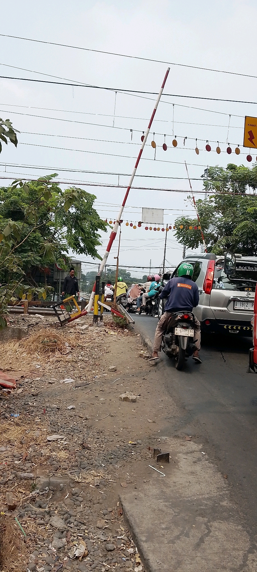 The image depicts a traffic scene at a railroad crossing in Bekasi, Indonesia. A red and white striped barrier arm is lowered, indicating an approaching train. Several motorcycles and a silver SUV are stopped in the lane, waiting for the train to pass. On the left side of the road, there is a person in a black shirt and green pants standing near some railroad tracks and debris. The area appears to be a busy intersection with overhead wires and decorative circular elements hanging from them. The road surface is asphalt, and the shoulders are unpaved with gravel and dry vegetation.