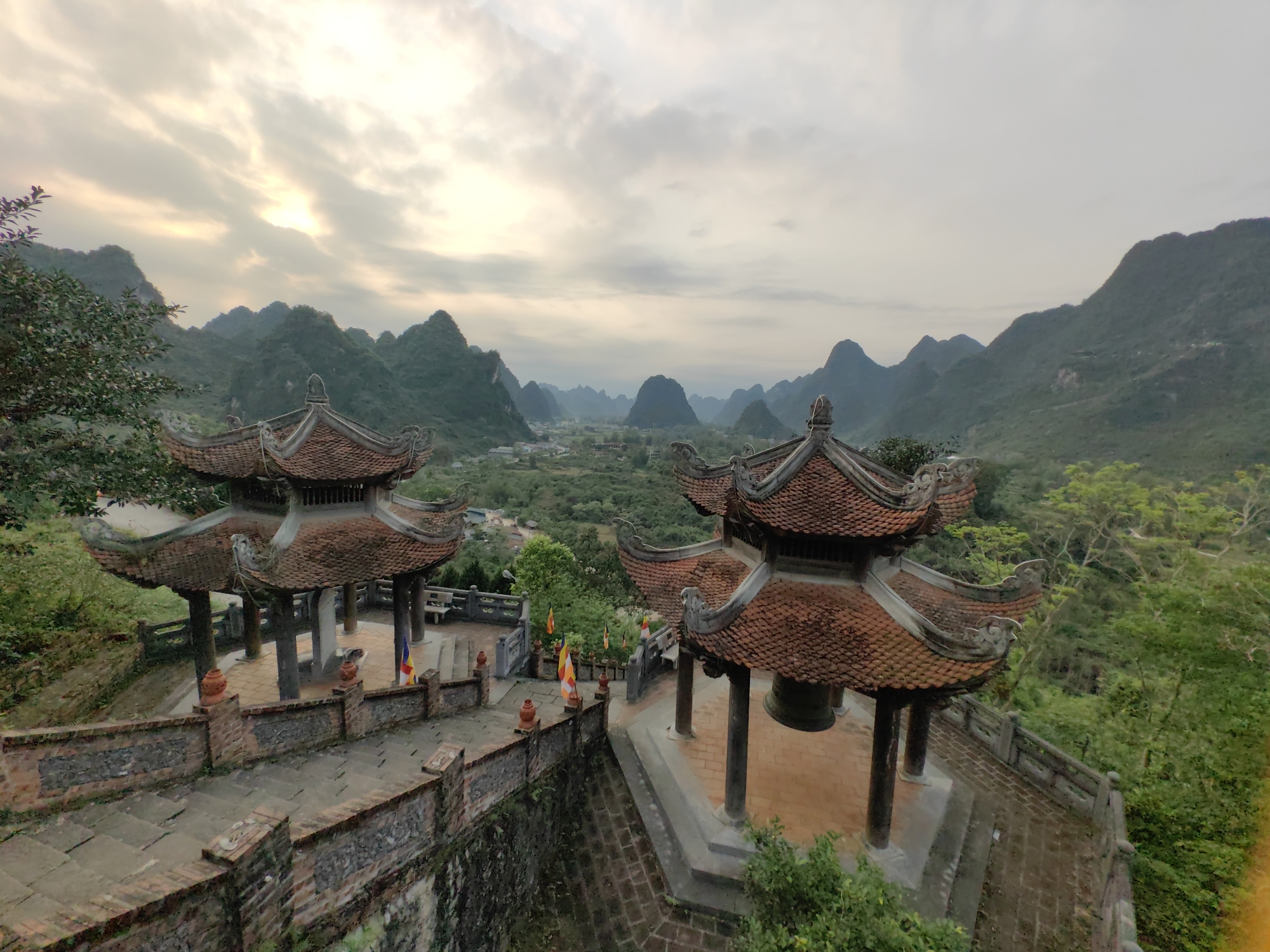 The image depicts a scenic view in Xã Đàm Thủy, Vietnam, featuring two traditional Vietnamese pavilions. Each pavilion has a multi-tiered, reddish-brown tile roof and is supported by dark wooden columns. The pavilions are connected by a stone pathway with brick walls and railings. Flags are displayed near one of the pavilions. In the foreground, there is a bell under a pavilion. The background showcases a landscape of green, forested hills and mountains under a cloudy sky, with a village nestled in the valley below. The lighting suggests either early morning or late afternoon.