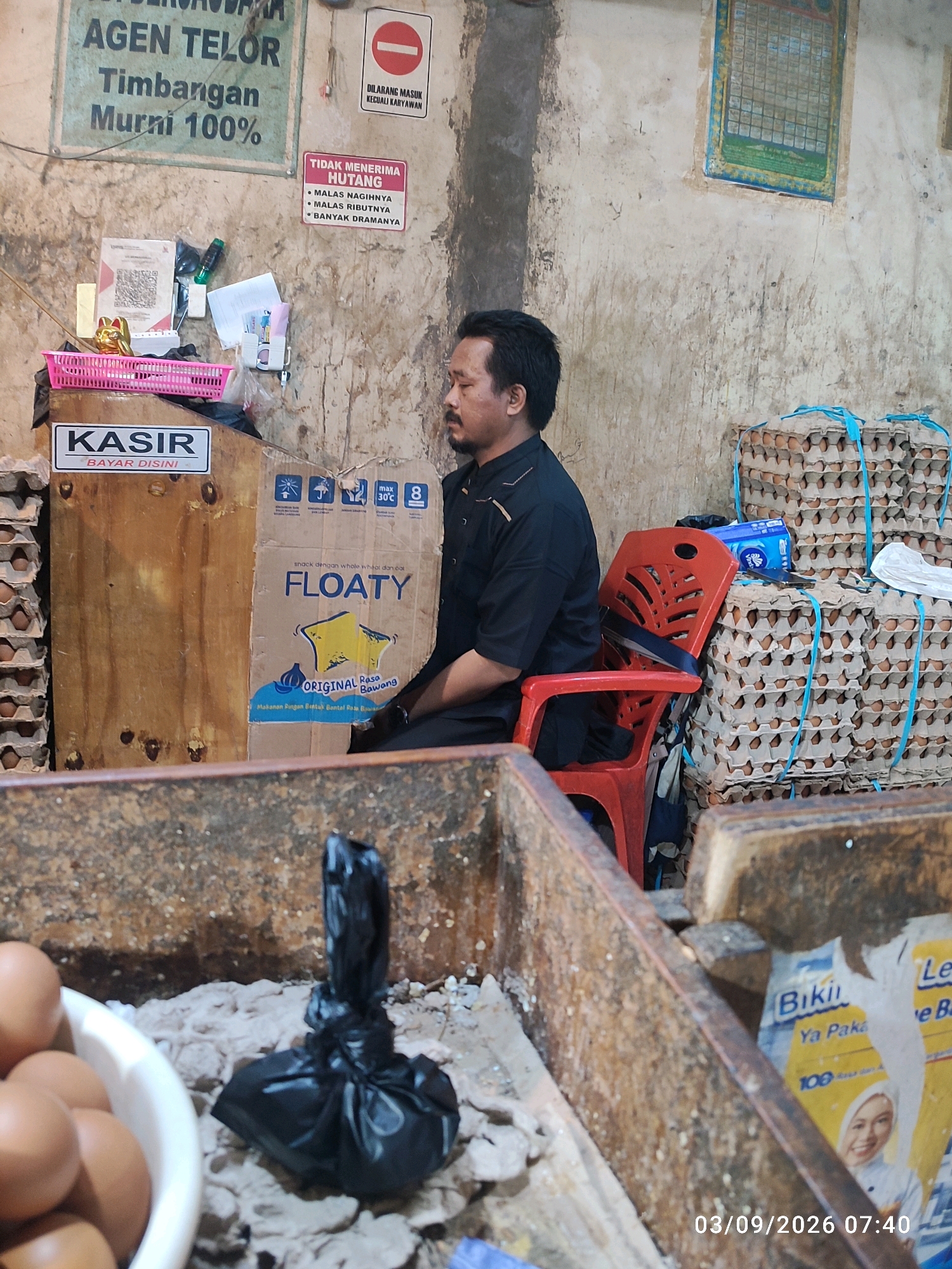 The image depicts a market stall in East Jakarta, Indonesia. A man in a black collared shirt is seated on a red plastic chair. Behind him are stacks of egg cartons, and to his left is a wooden counter labeled "KASIR BAYAR DISINI" (Cashier Pay Here). A cardboard box with the word "FLOATY" is also visible on the counter. The stall is cluttered with various items, including a pink basket, a QR code for payment, and a small golden cat figurine. In the foreground, a white bowl filled with eggs and a black plastic bag are on a rough surface. The background shows a textured, possibly concrete wall with some signs, including one indicating "DILARANG MASUK KECUALI KARYAWAN" (No Entry Except Employees). The overall scene suggests a busy, functional market environment.